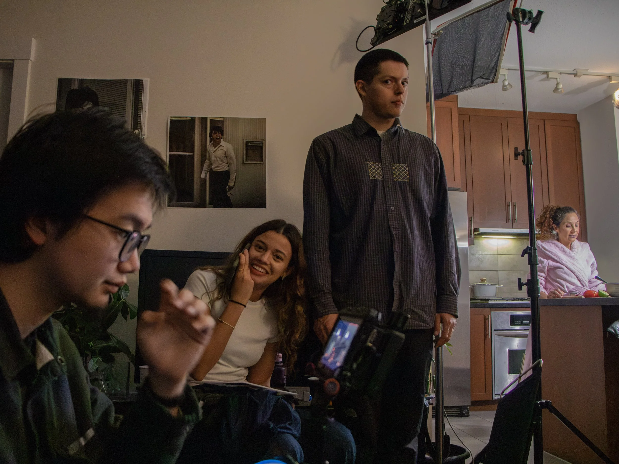 A group of people gathered indoors at a table, with one person standing near film equipment, and others engaged with their phones or smiling. There is a picture of a woman in a white shirt on the wall behind them, and the scene appears to be a casual