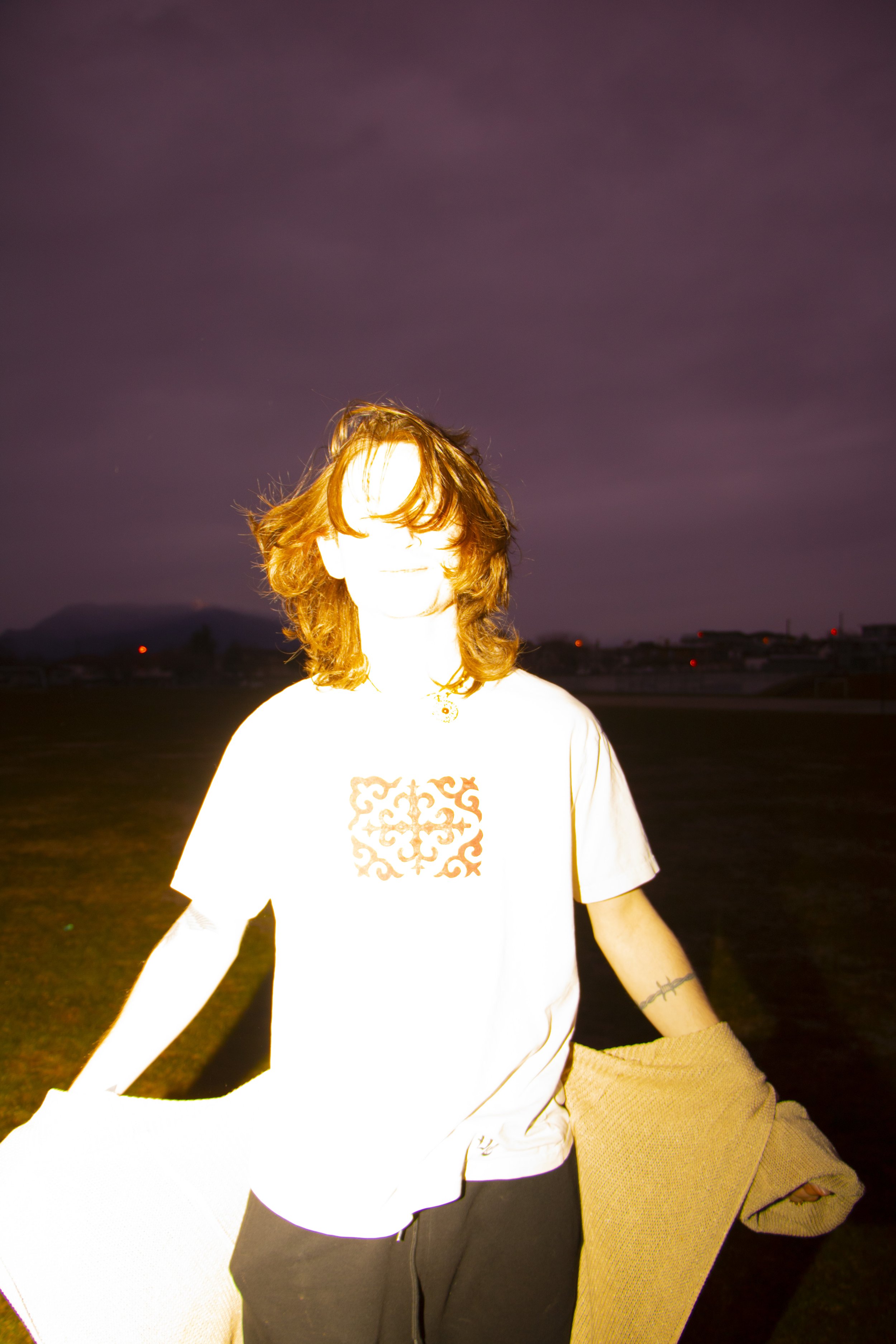 Person with shoulder-length hair standing outdoors in a dark nighttime sky, wearing a white T-shirt with an intricate design on the front, and holding a beige coat or blanket.