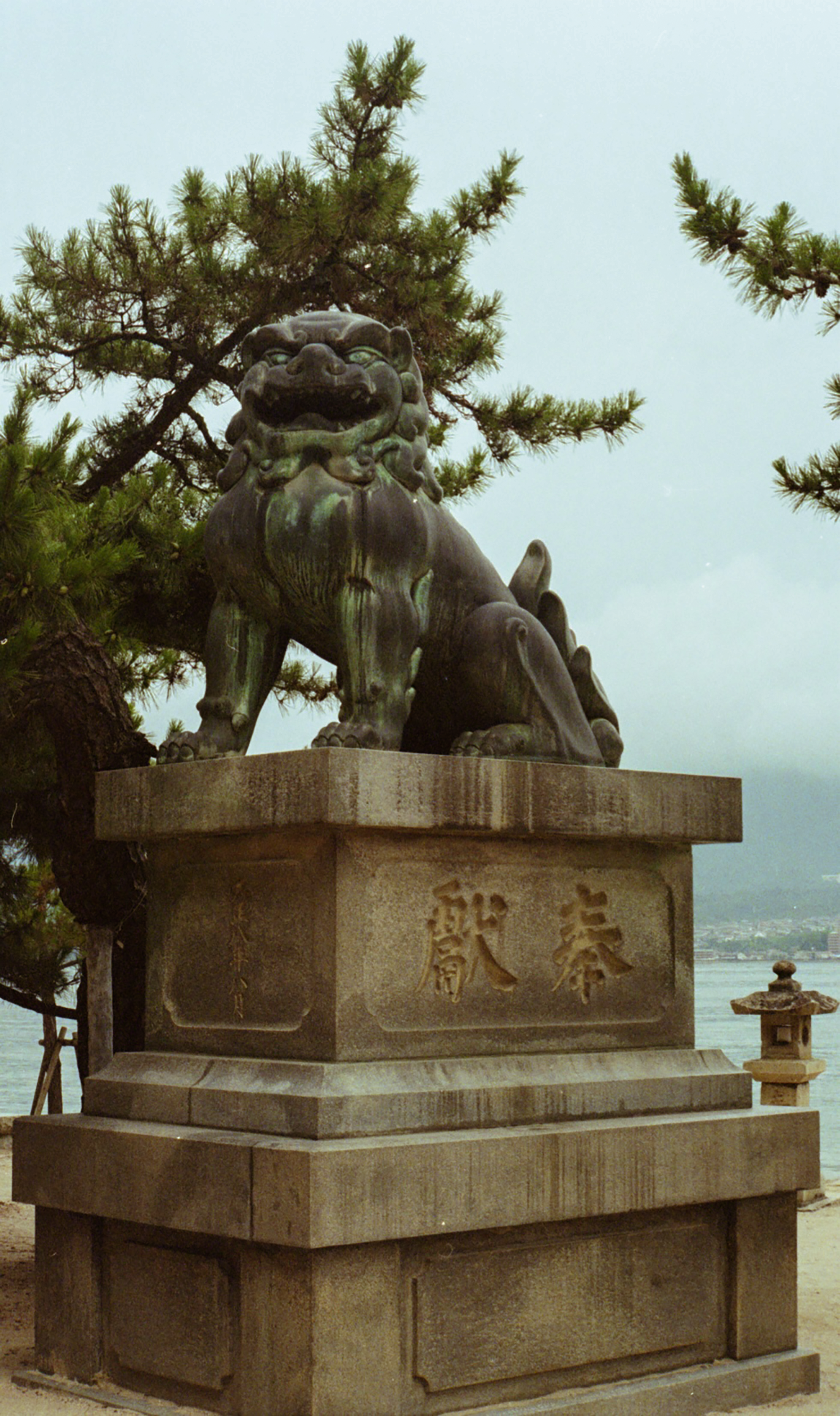A traditional Japanese lion-dog statue, called a Komainu, sits atop a stone pedestal with Japanese characters engraved on it, surrounded by pine trees and overlooking a body of water.