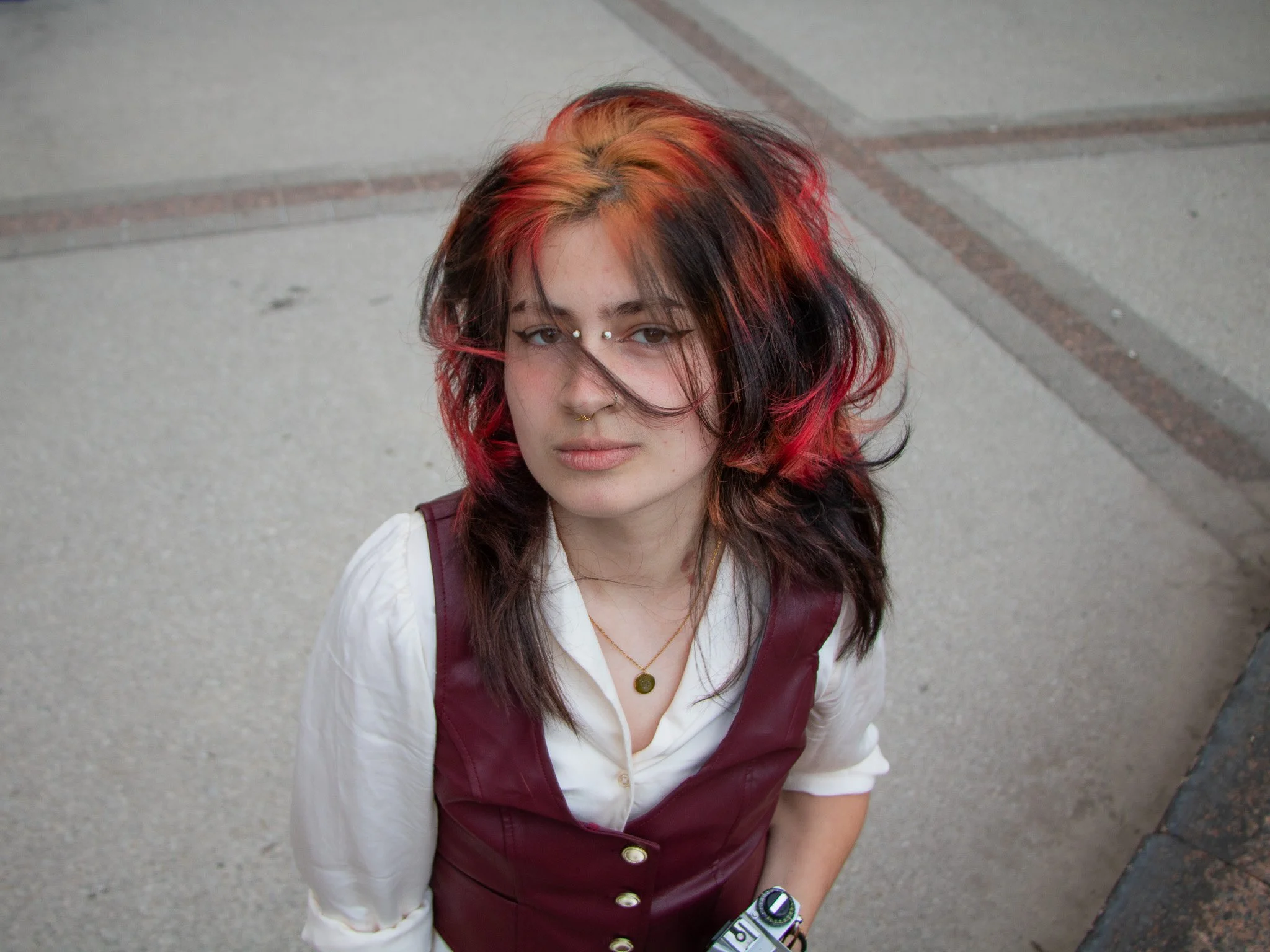 A young man with multicolored hair in shades of red, orange, and brown, looking up at the camera with a neutral expression, standing on a paved sidewalk.