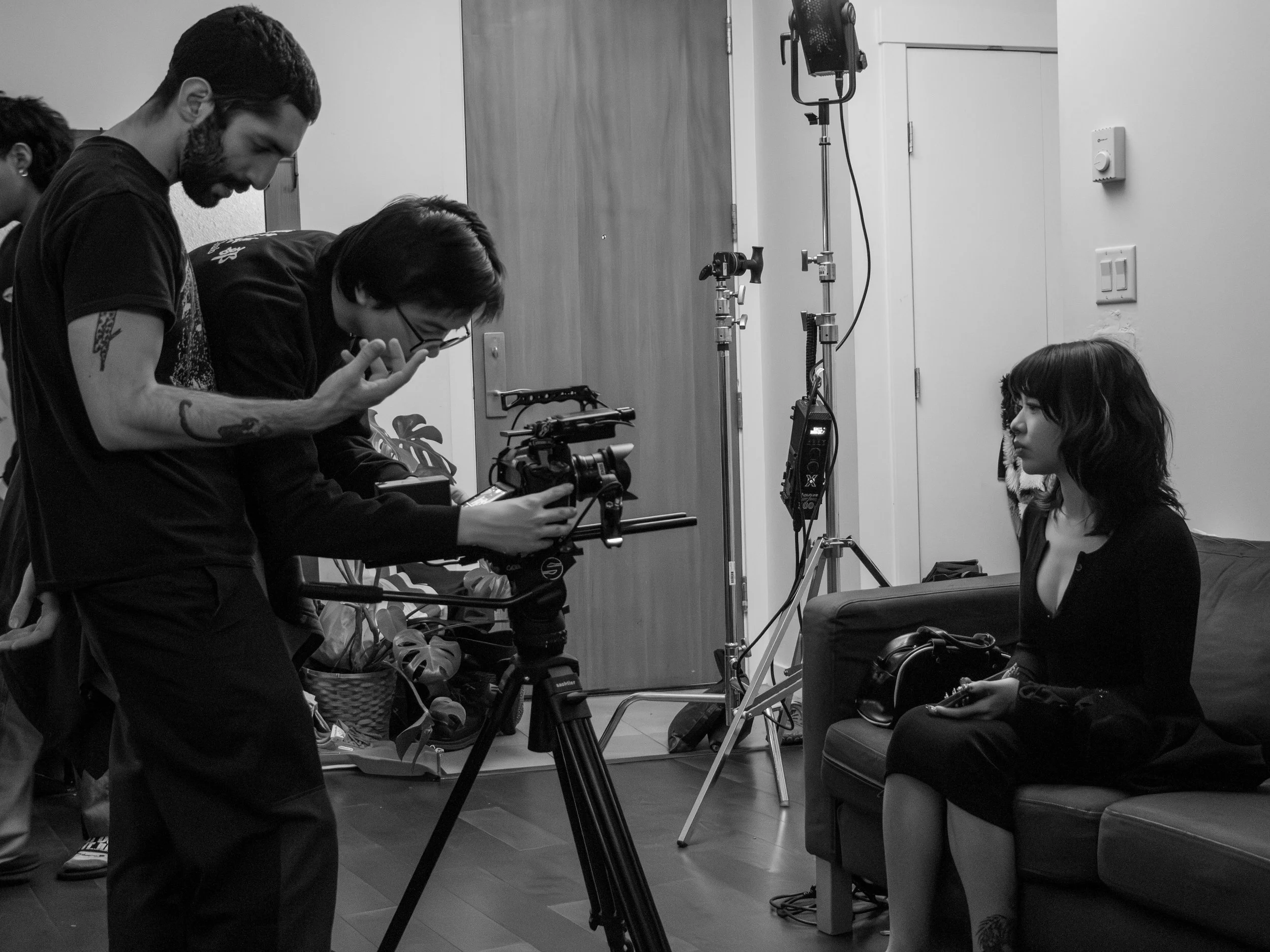 Three people in a room, with one person sitting on a chair and two people standing nearby. The scene appears to be a film or photo shoot setup, with equipment such as lights and a camera present.