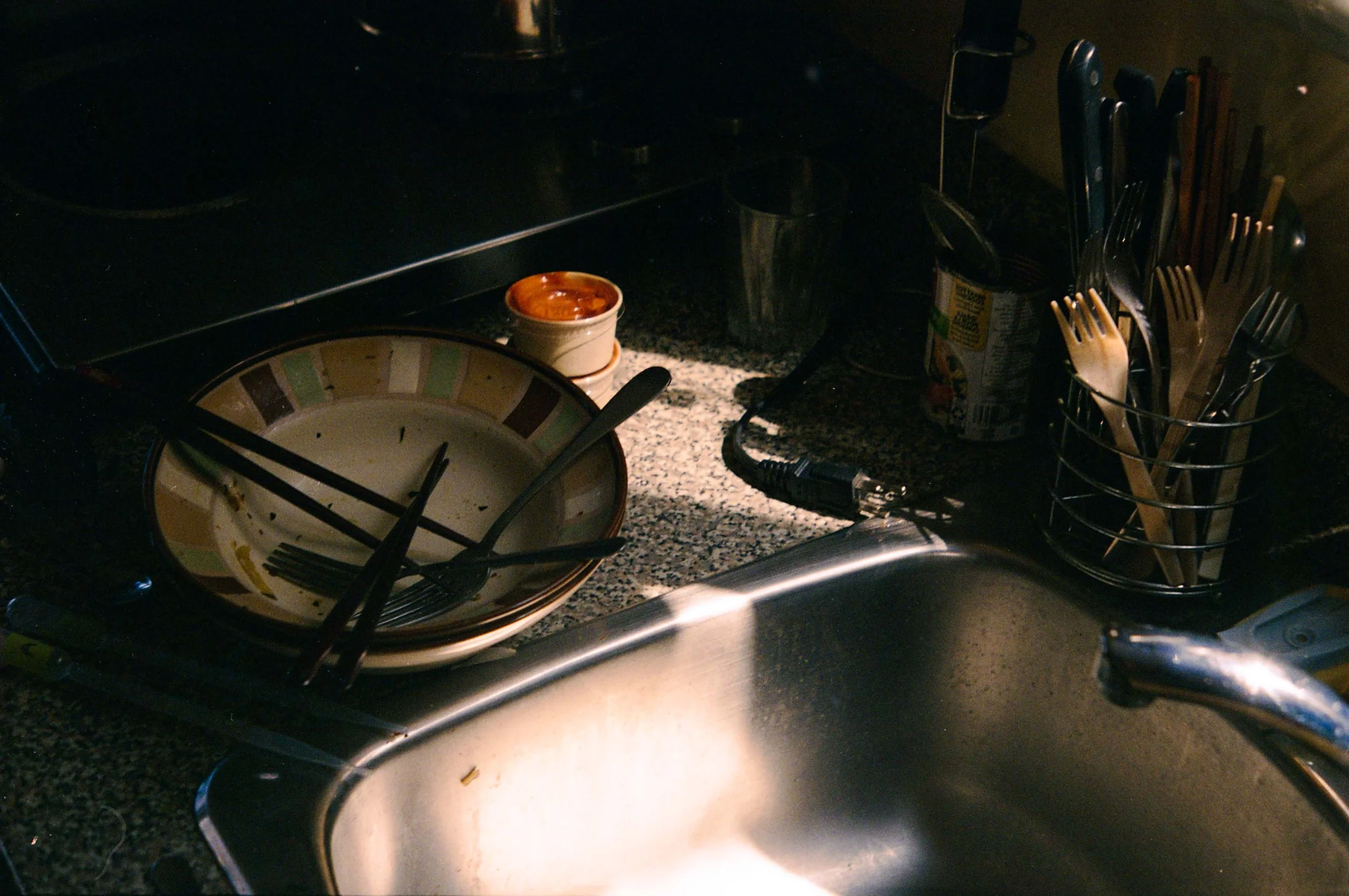 Kitchen sink with dirty dishes, a small bowl, and utensils on a dish rack on a dark countertop.