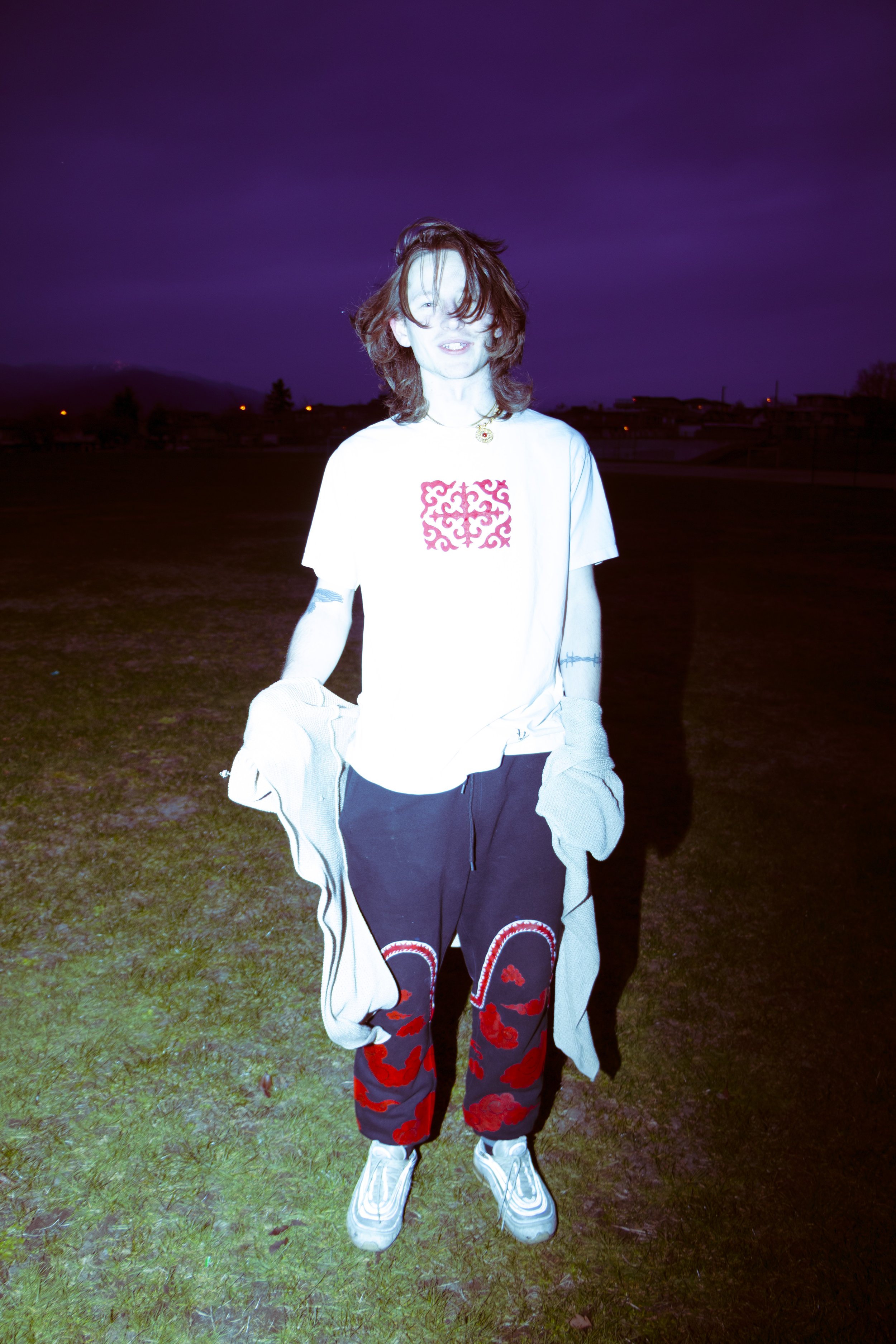 Person standing outdoors on grass at night, wearing a white T-shirt with red kyrgyz design, patterned pants, sneakers, and holding a gray cardigan, with a dark purple sky and distant city lights in the background.