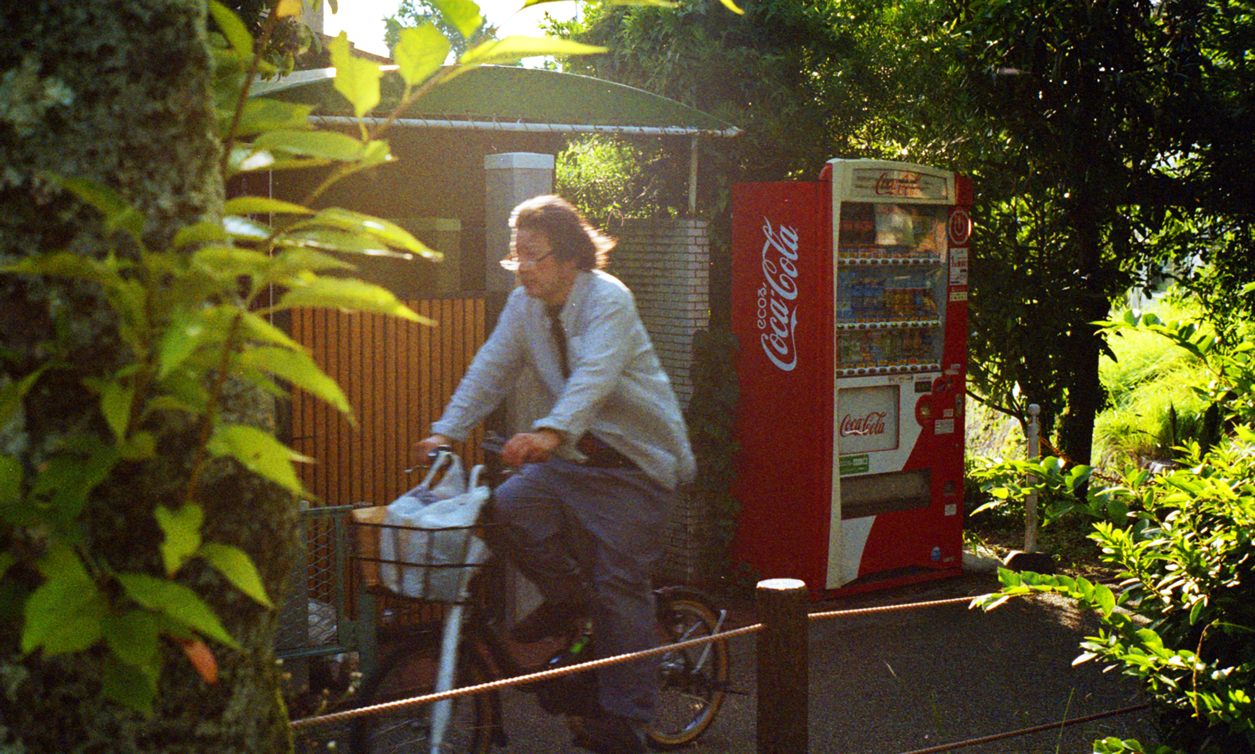 A man riding a bicycle past a red Coca-Cola vending machine in a green, leafy outdoor setting.