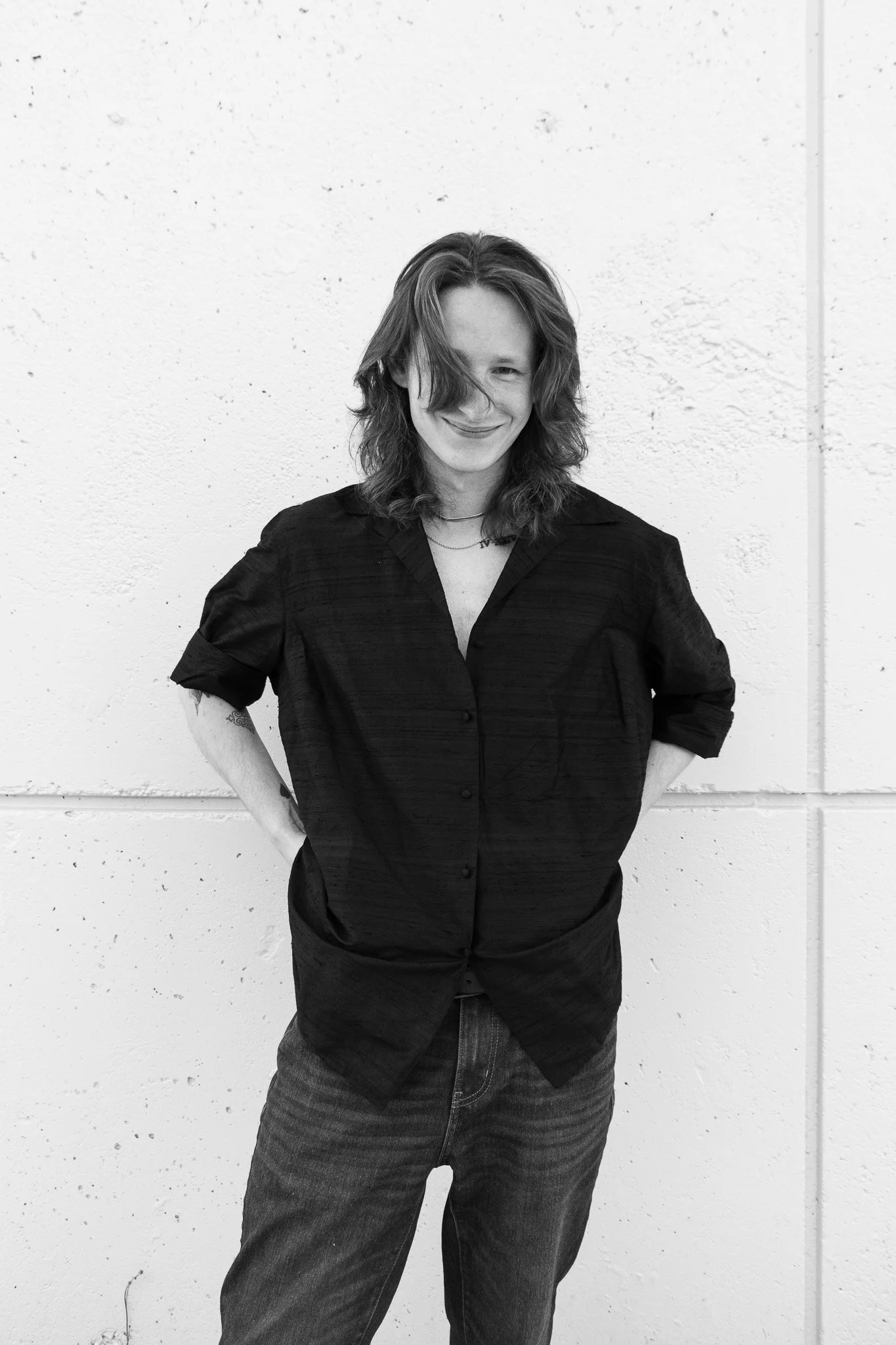 Black and white photo of a young man with wavy hair, smiling, standing with hands on hips against a textured wall.