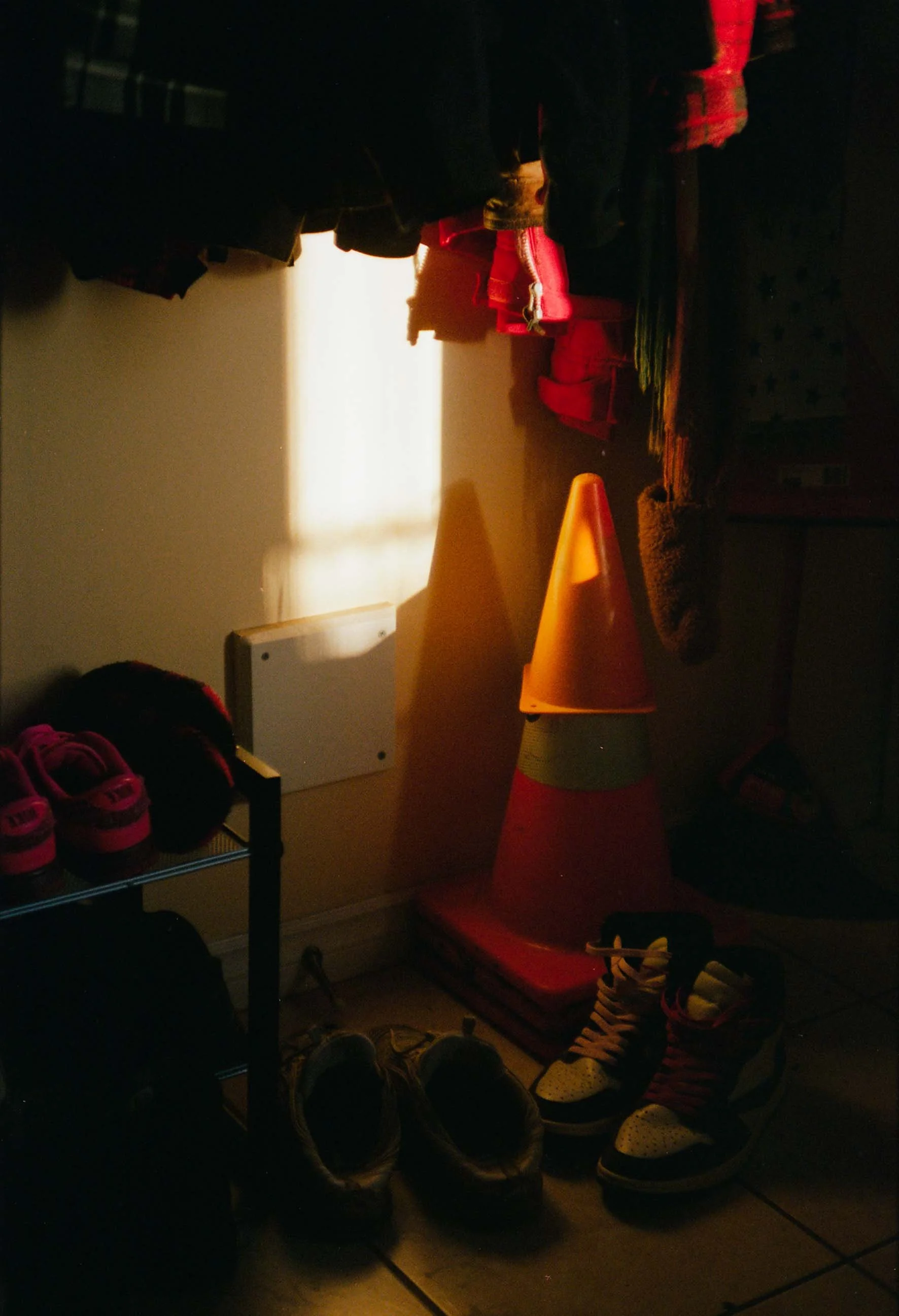 A dimly lit room with wall-mounted coat hooks holding jackets and coats, shoes including pink and yellow sneakers, a corner with stacked storage bins, and orange traffic cones