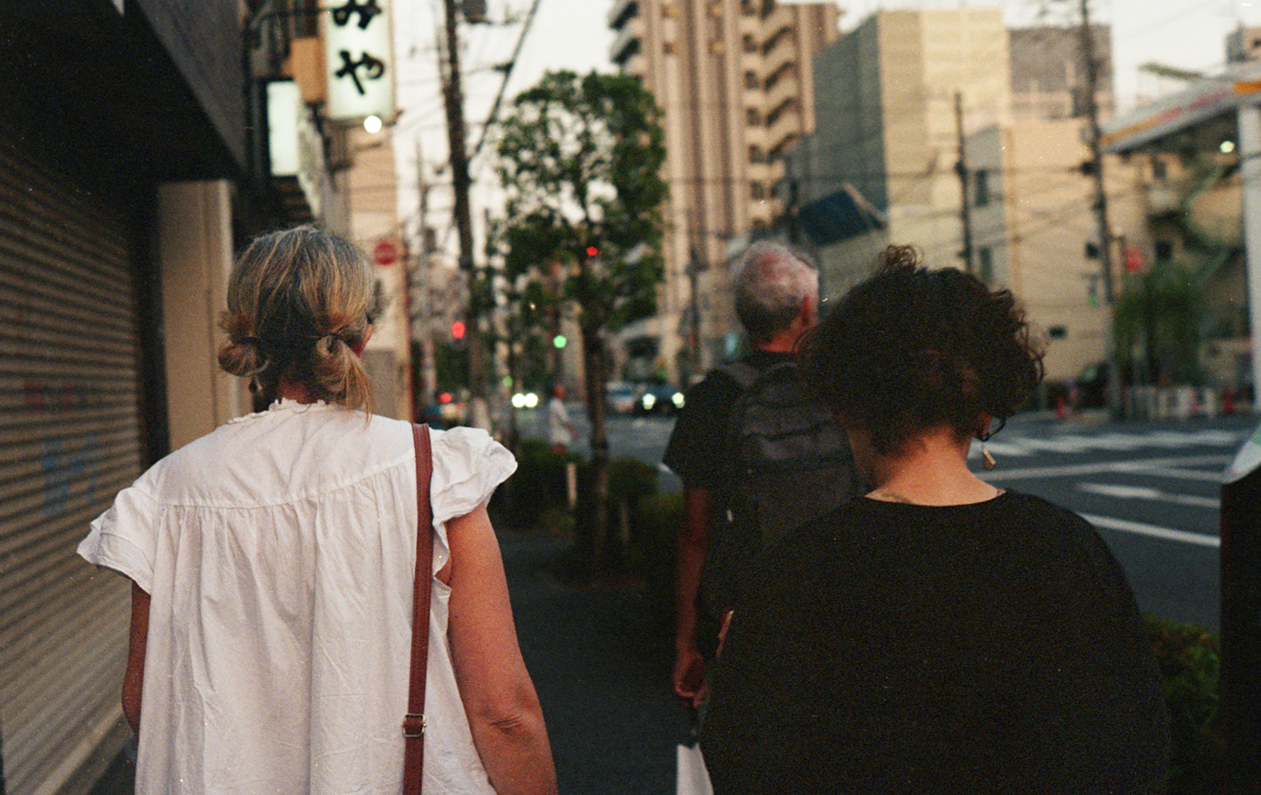 Two women walking along a city sidewalk during the evening, seen from behind. City buildings and a street with traffic are visible in the background.