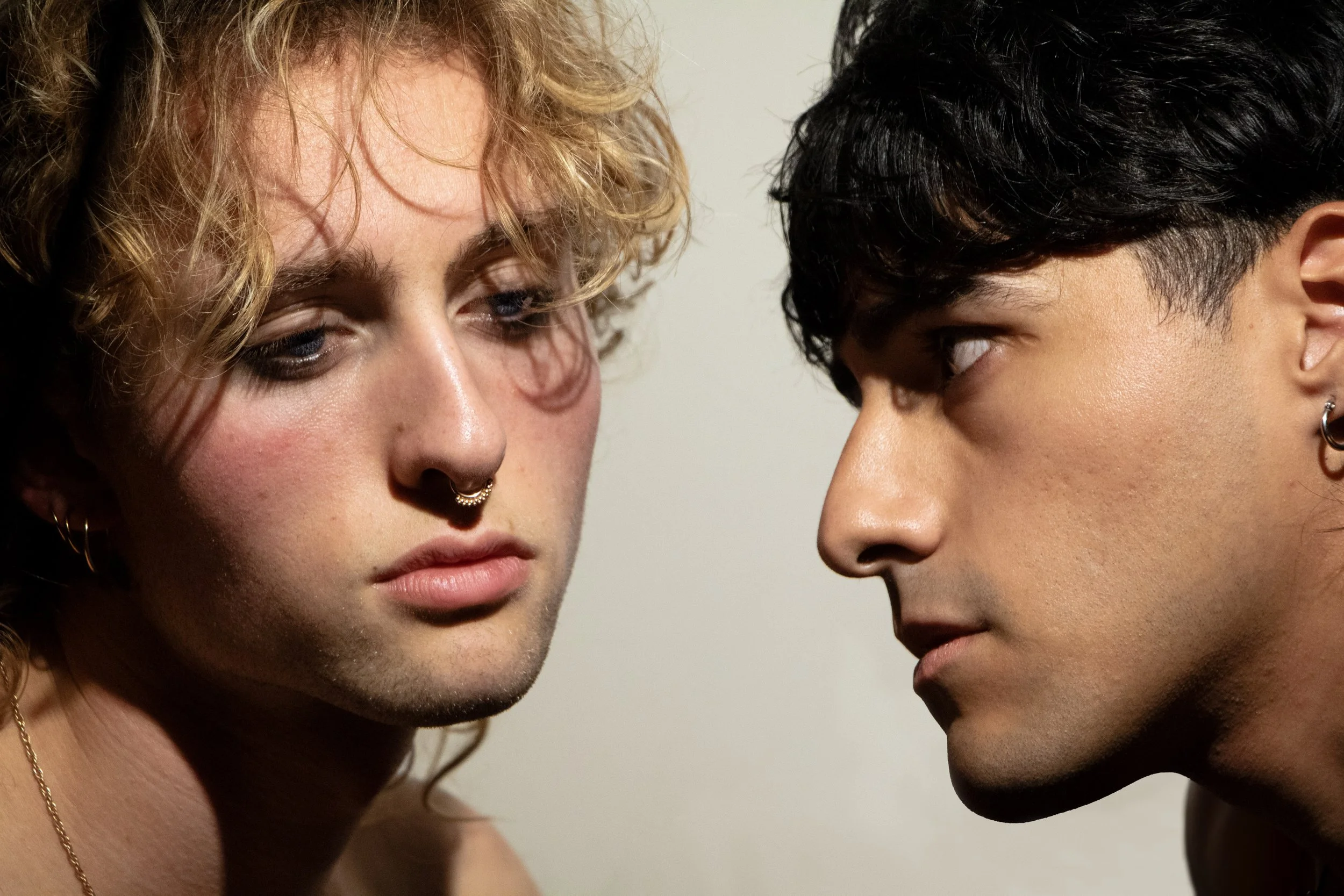 A close-up photo of two with their faces close The person on the left has curly blonde hair, light skin, and is wearing a nose ring, with a neutral expression. The person on the right has short, curly dark hair, medium skin tone, and is also looking at the other person with a neutral expression.
