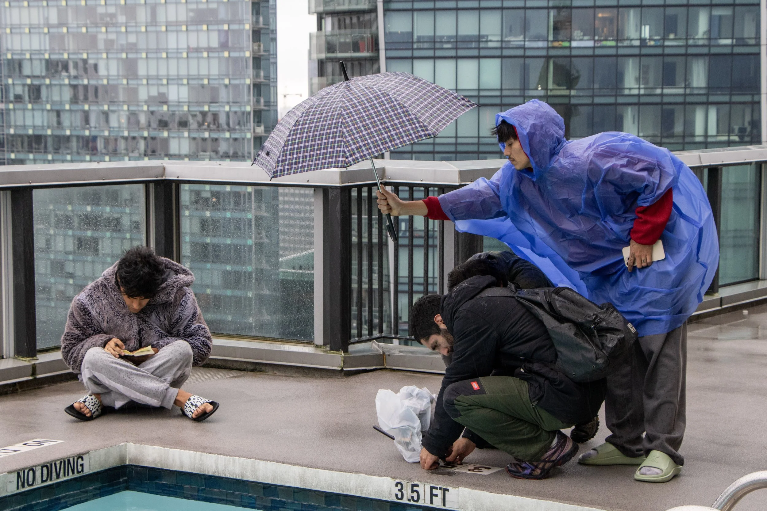 A group of four people on an outdoor rooftop in cityscape, with three crouching and one sitting, all wearing rain gear or casual clothing, one holding an umbrella, others focused on a task near a swimming pool with a 'No Diving' sign.