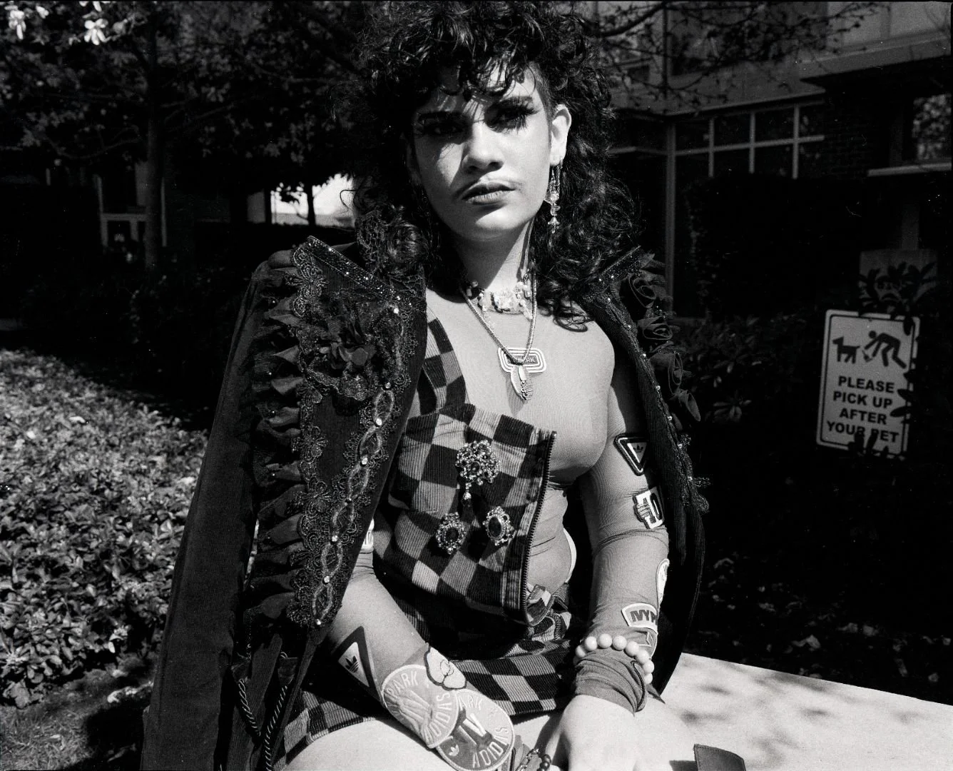 A queer person with curly hair, wearing a patterned jacket adorned with embellishments, sitting outdoors in black and white. They are accessorized with layered necklaces and earrings, with a serious expression on their face. 
