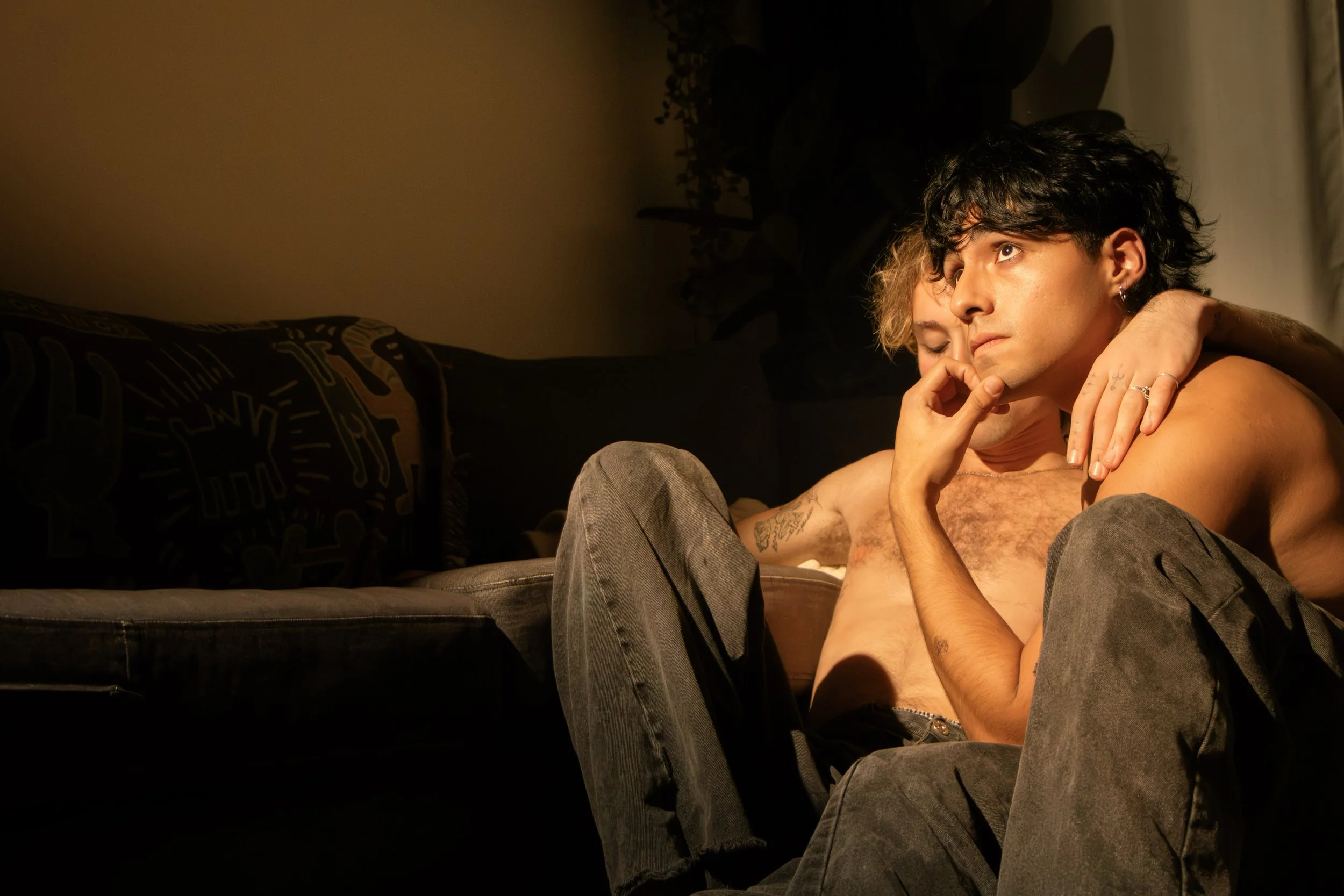 A young man with curly dark hair and a second person with blonde hair hugging each other on a couch in a dimly lit room.