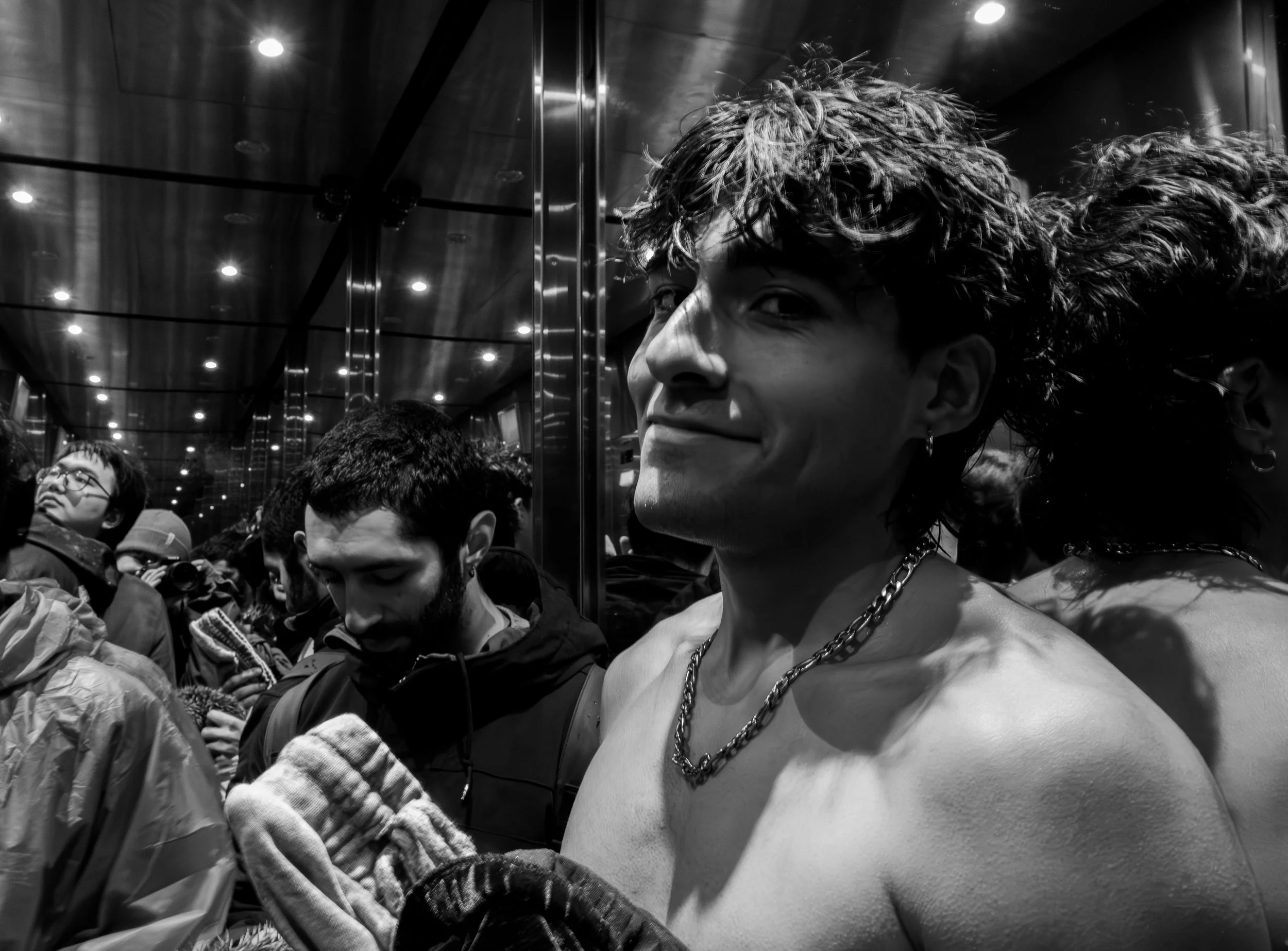 A young person with curly hair and chains around their neck in an elevator, smiling, surrounded by other people.