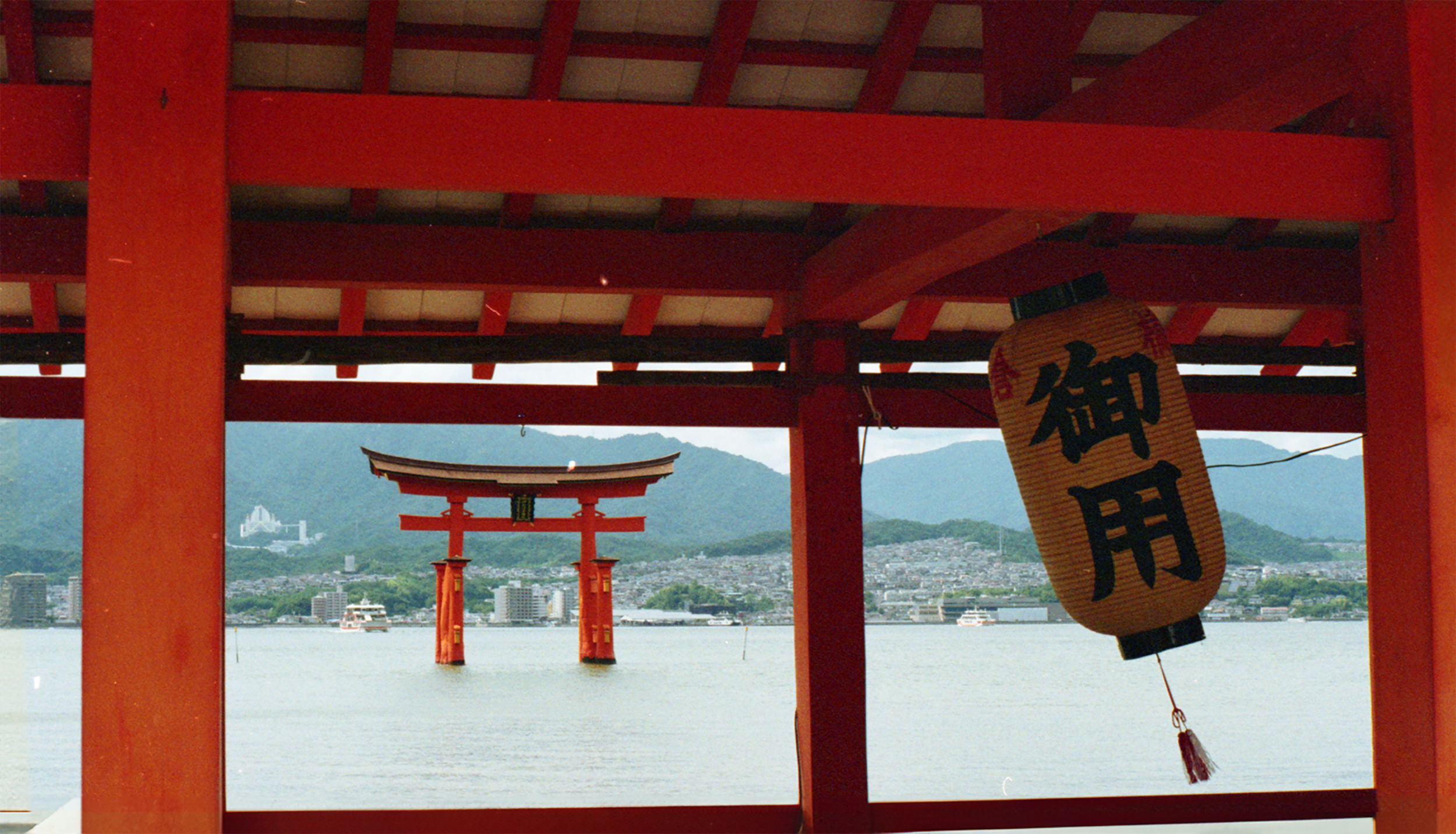 Red torii gates leading toward a view of a city and water with mountains in the background, with a hanging Japanese lantern with black kanji characters