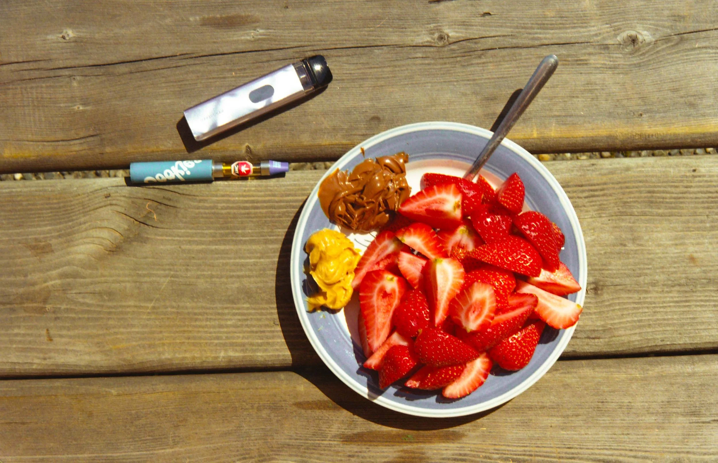 A plate of sliced strawberries with nutella and peanut butter on a wooden bench with a vape and a cannabis pen nearby.