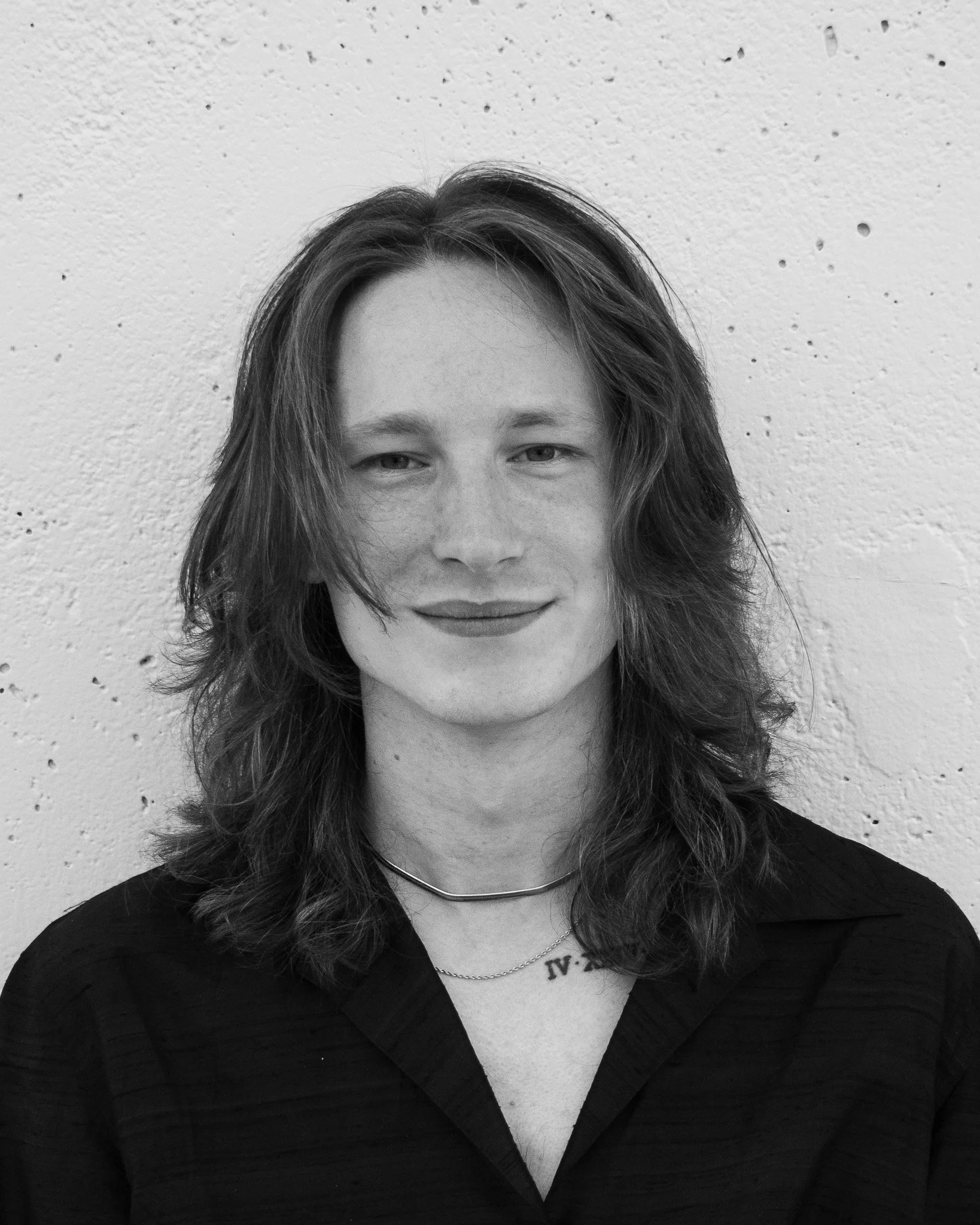A black and white photo of a young man with shoulder-length wavy hair, wearing a necklace and a V-neck top, against a textured wall background.