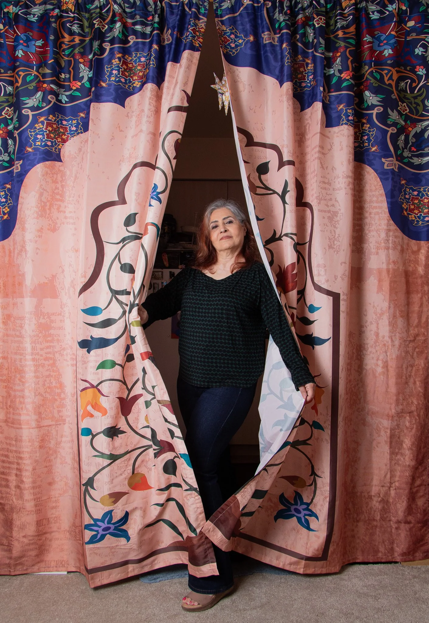 A woman with gray hair standing behind a partially open, patterned curtain, wearing a black top and dark jeans.