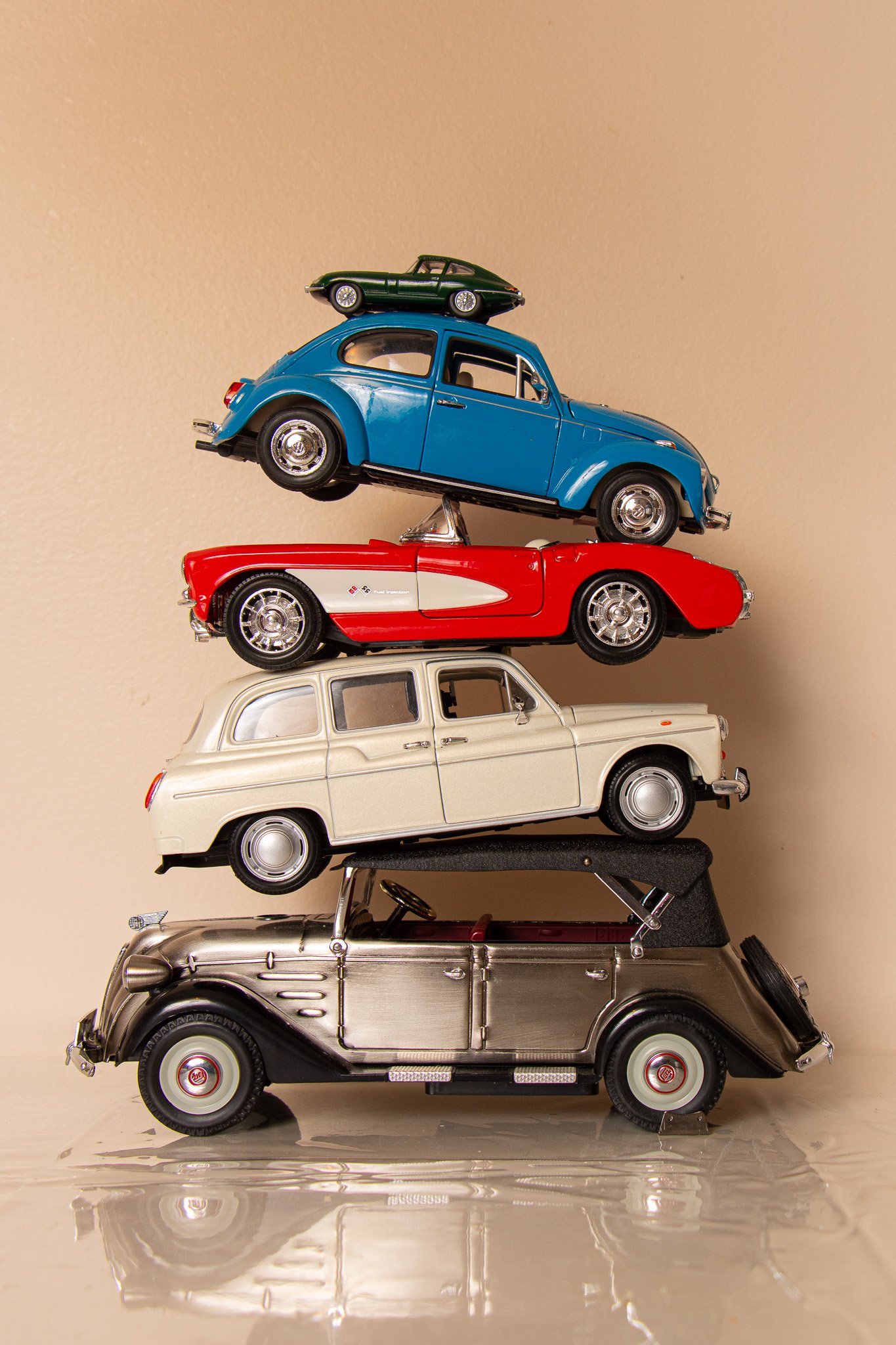 Stacked models of classic cars, with a black vintage car at the bottom, a silver vintage car above it, a red toy sports car, a blue vintage car, a green sports car, all on a plain background.