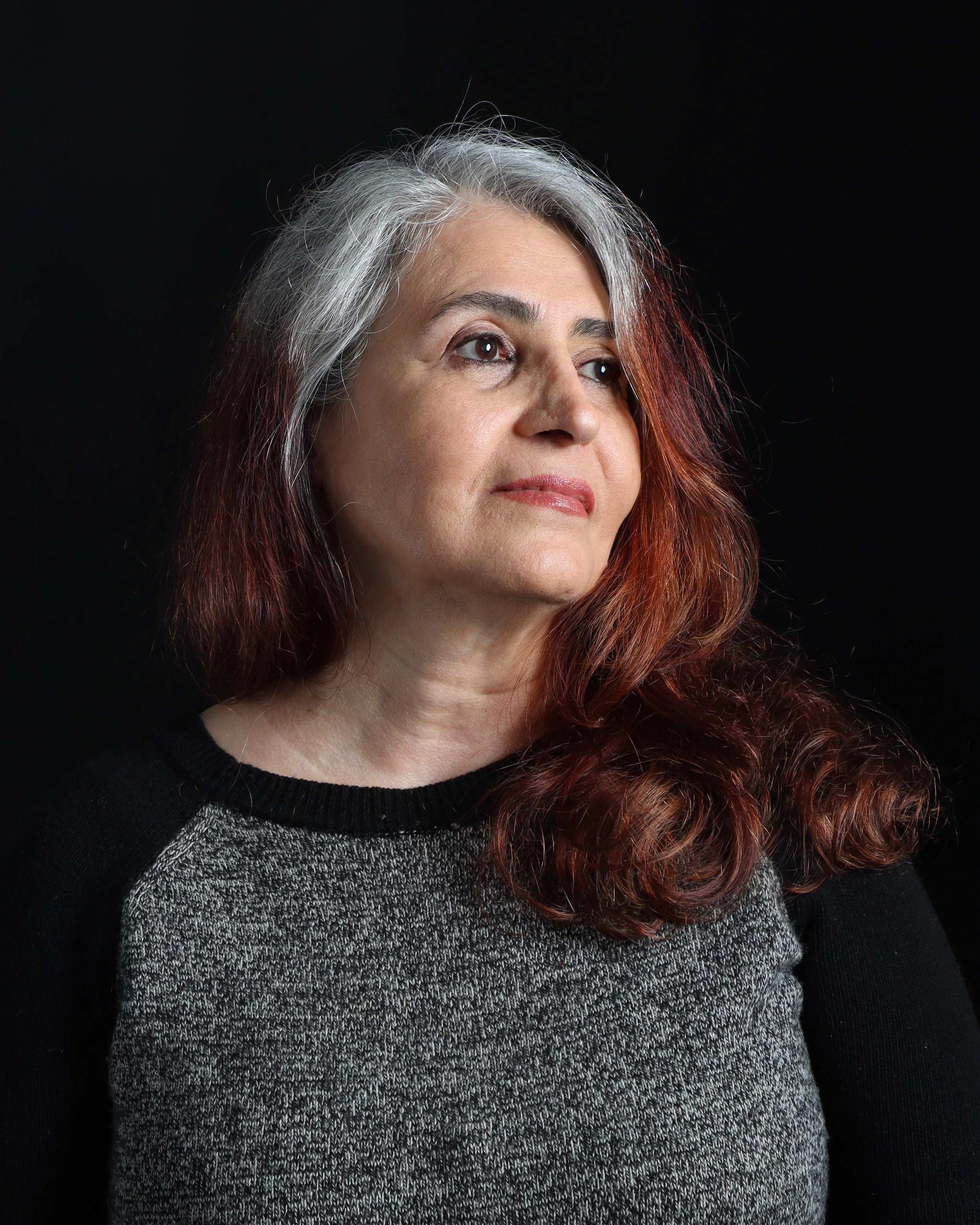 A woman with long gray and red hair, wearing a black top and a gray sweater, looking slightly upwards against a solid black background.