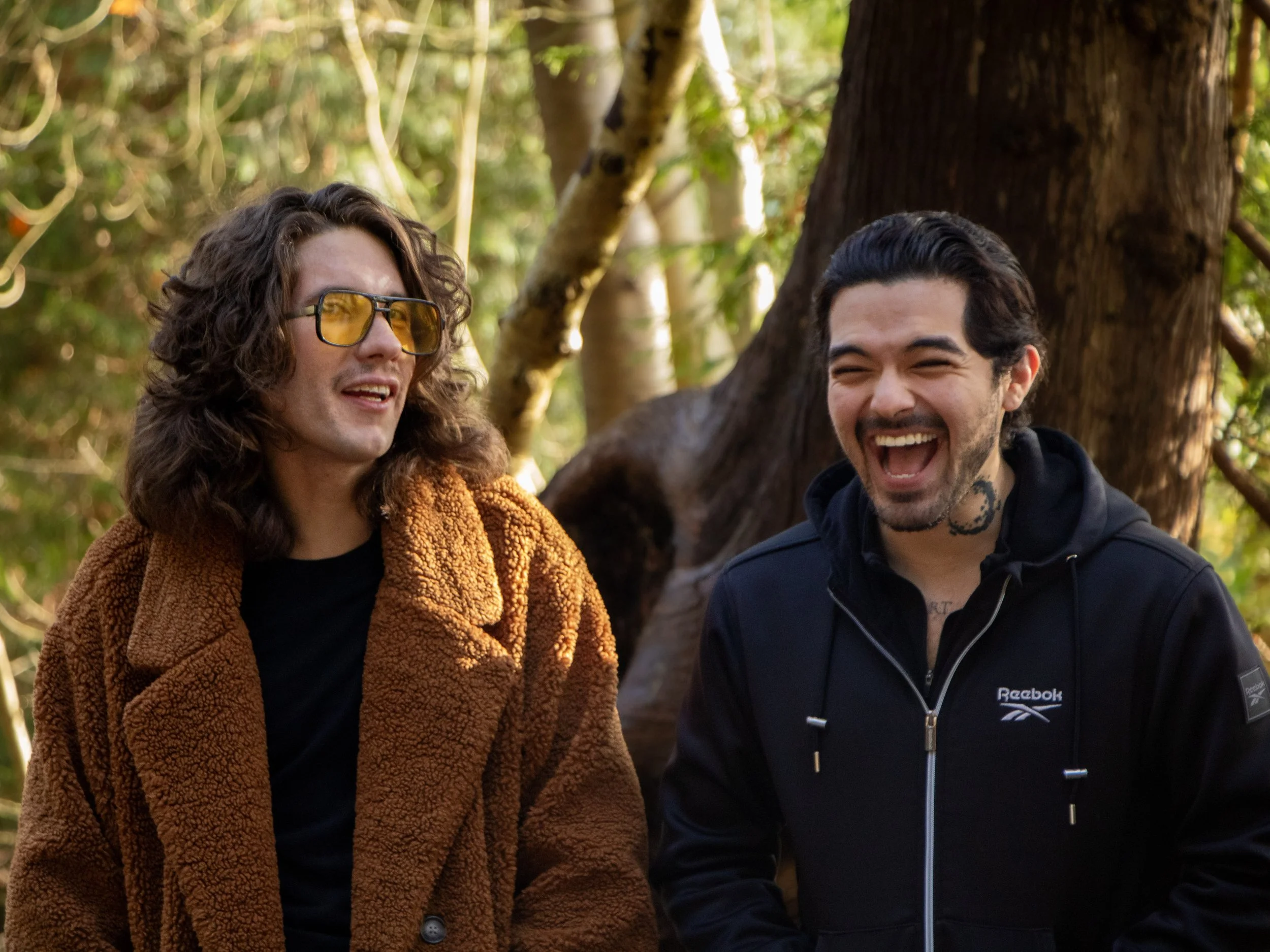 Two men are laughing and enjoy each other's company outdoors in a wooded area. One man wears a brown fuzzy jacket and yellow-tinted glasses, and the other wears a black Reebok jacket.