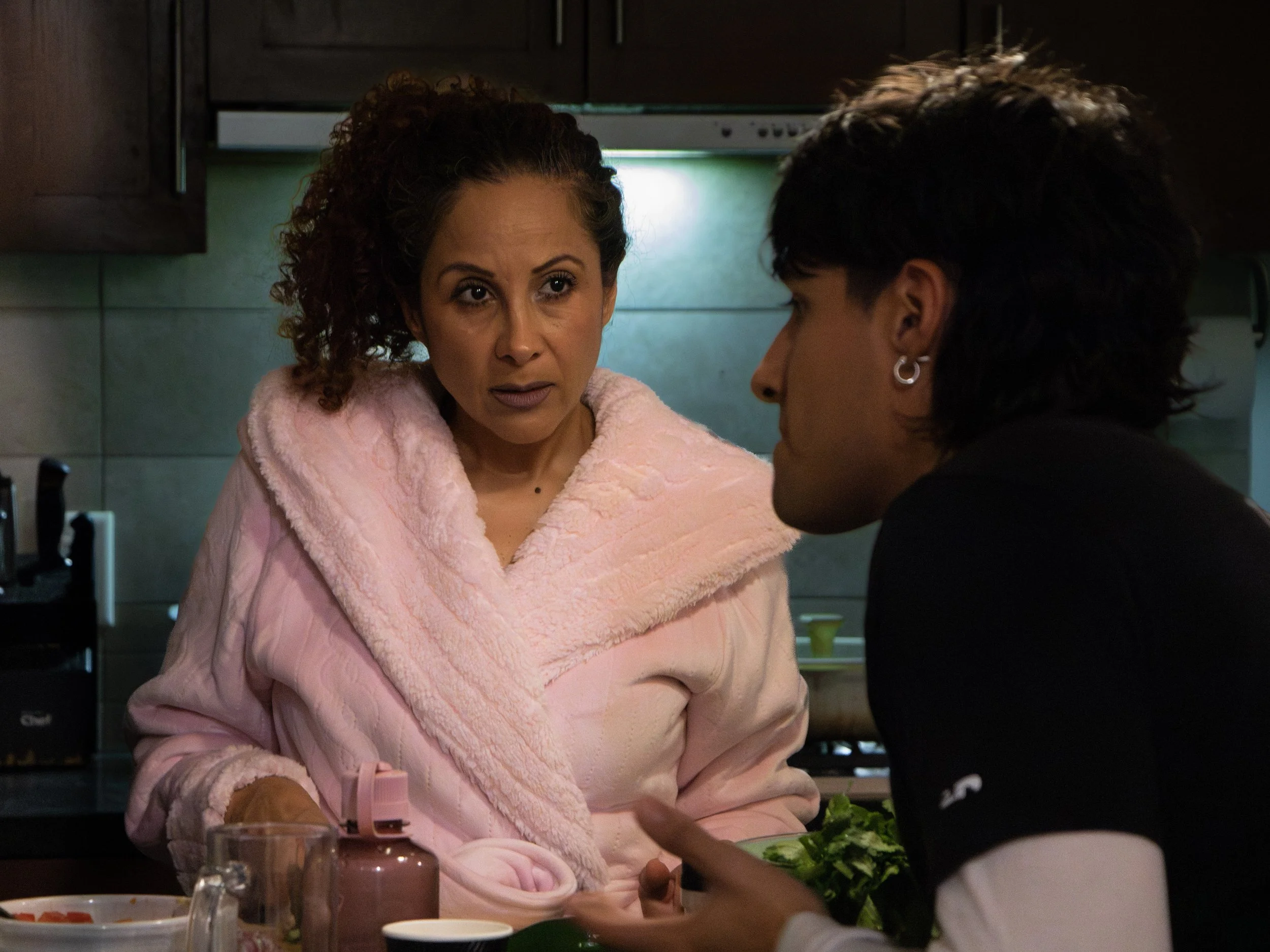 Two women are in a kitchen, one wearing a pink fuzzy coat, and the other wearing a dark shirt. They are preparing food together, with a salad bowl visible on the counter. The woman in pink has curly hair and is looking at the other woman, who has dar