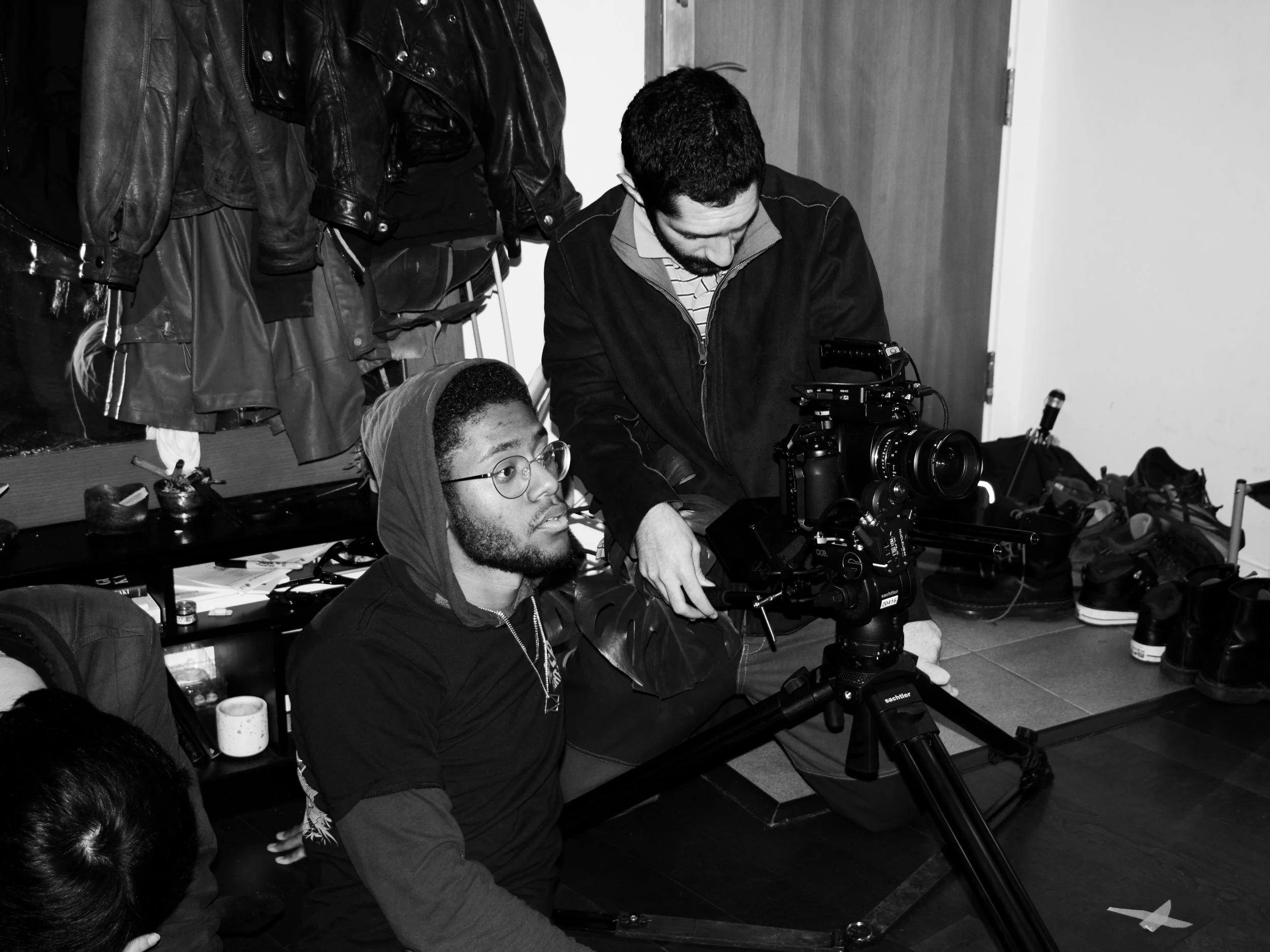 Two men working with professional camera equipment in a room, one seated and the other standing, surrounded by various items and jackets hanging on hooks.