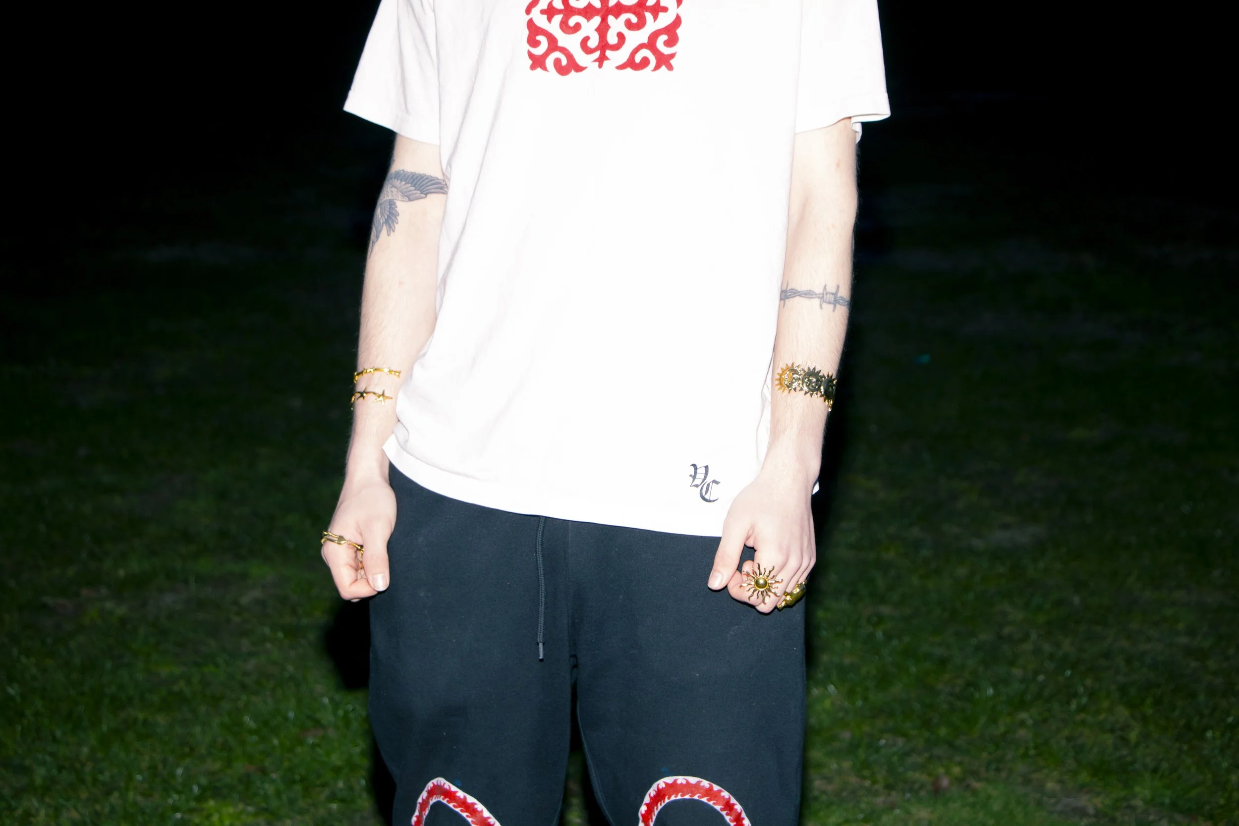 A person wearing a white t-shirt with a red kyrgyz design on the chest and black pants. They have tattoos on their right arm, jewelry including bracelets and rings, and are standing outdoors on a grassy area at night.
