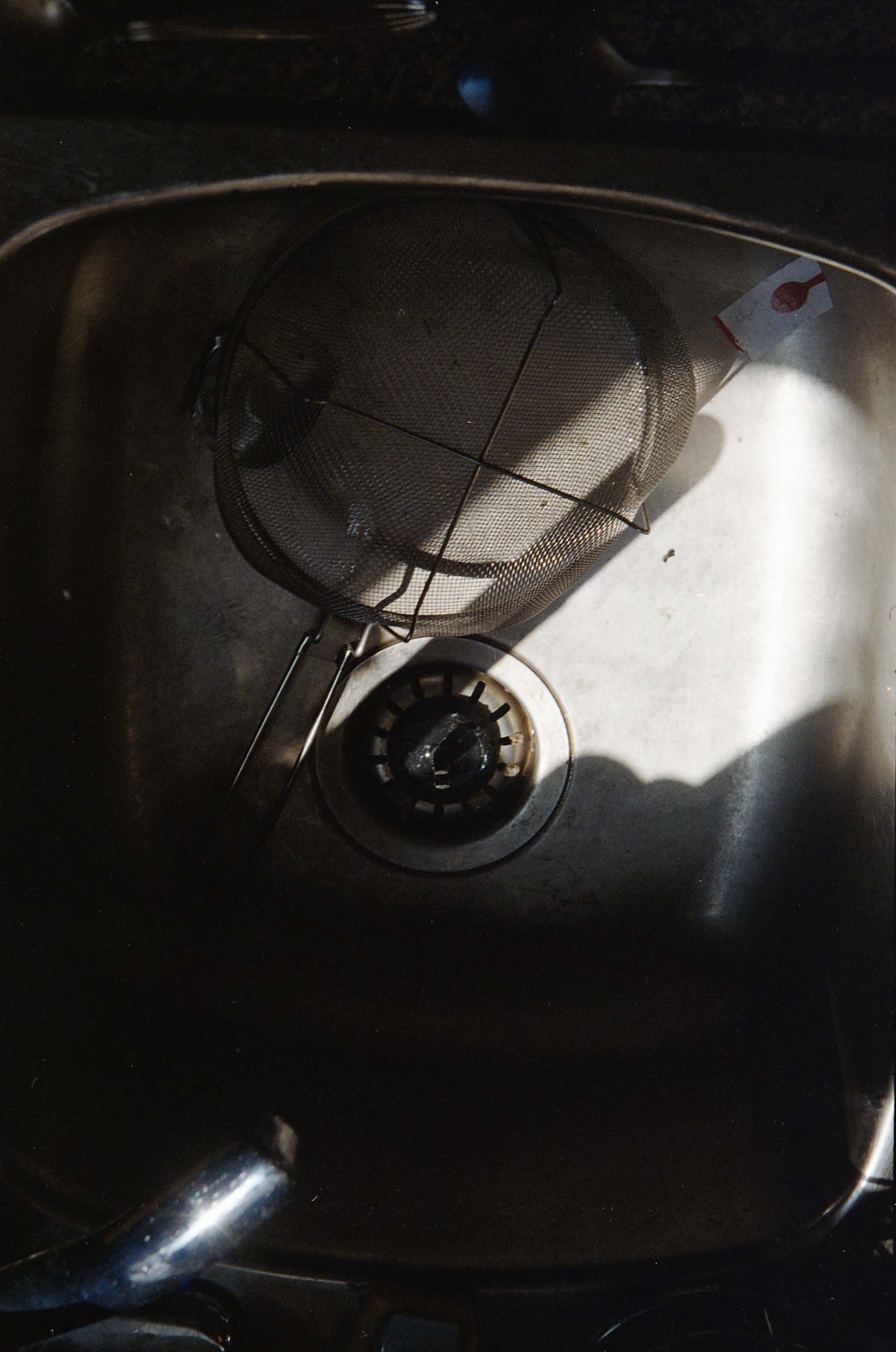 Inside of a kitchen sink with a metal strainer and a utensil, possibly a spoon, resting in the drain.