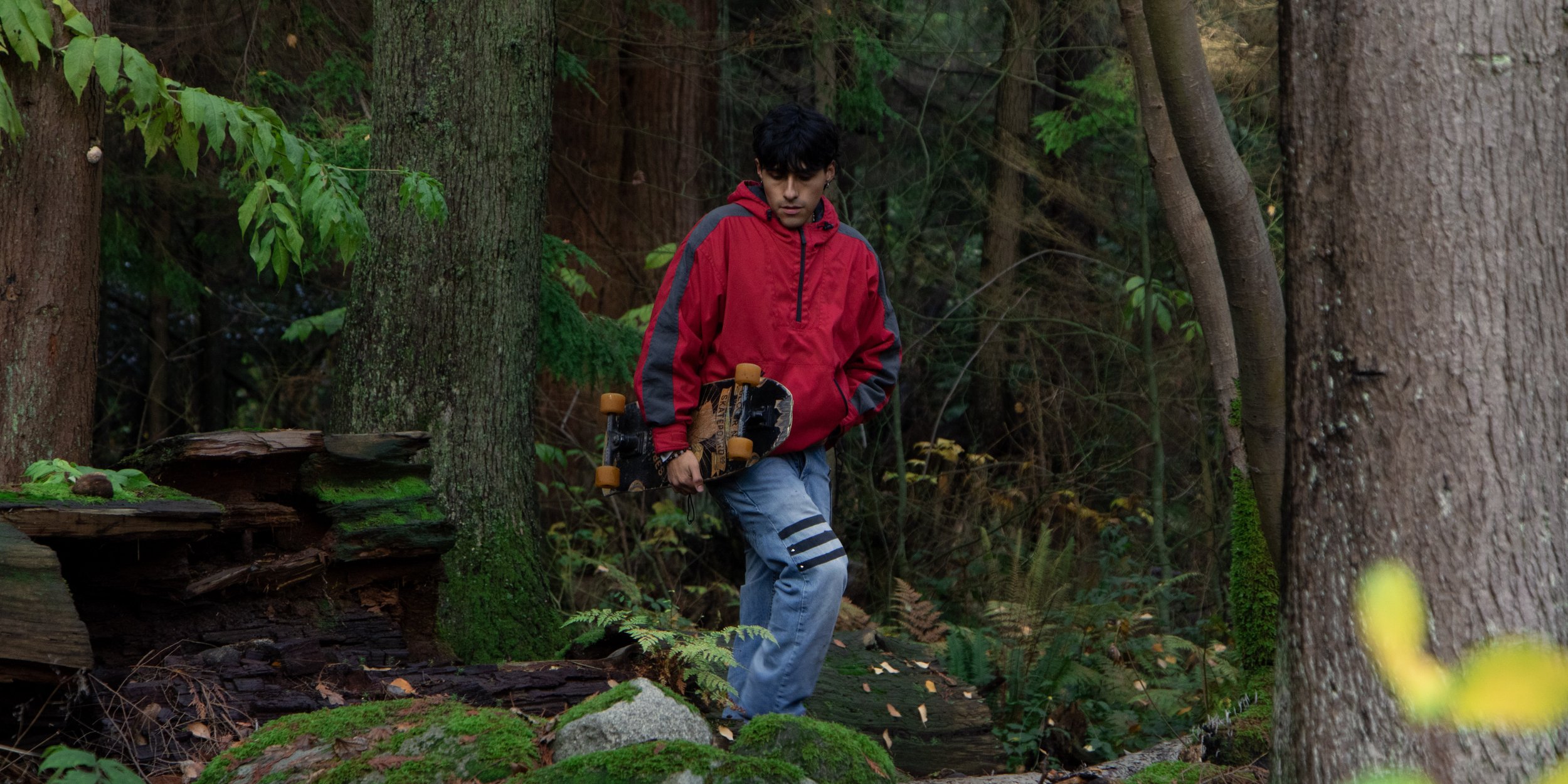 A young man in a red and black jacket holding a skateboard, walking through a dense green forest.