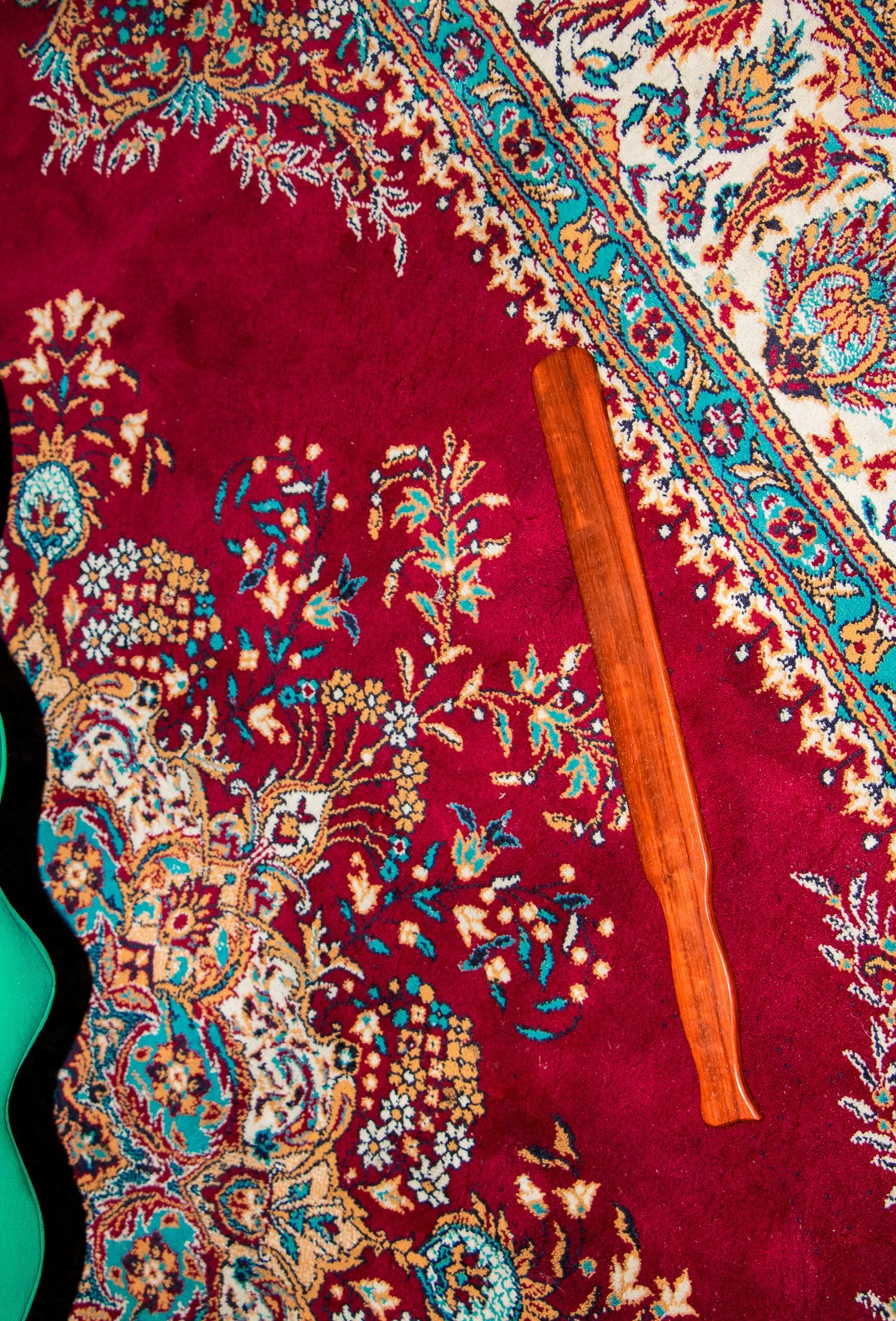 Close-up of a decorative Persian-style rug with intricate floral and geometric patterns in red, blue, beige, and gold, with a wooden spanker resting on top of the rug.