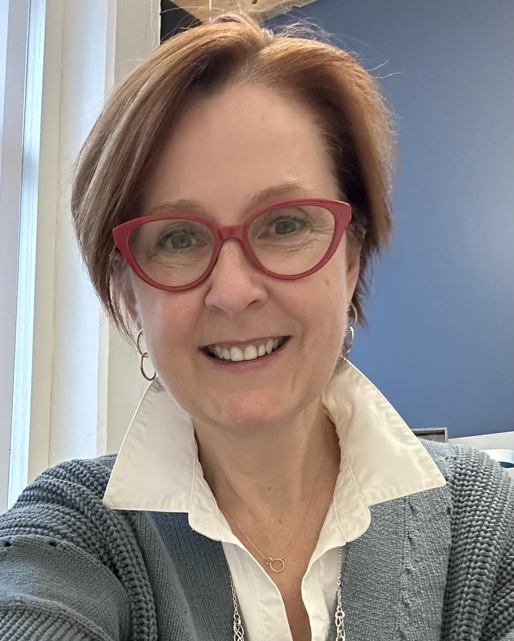 headshot of laura flynn, ceo of laura flynn coaching, sporting a beautiful red hair bob cut and red glasses and a muted dark teal knit sweater over a collar-popped white button down