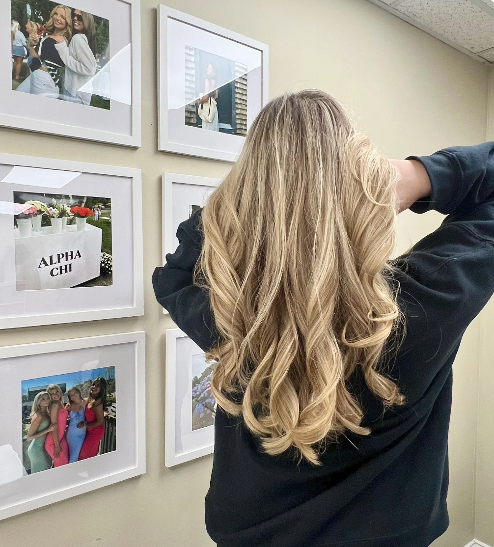 A woman with long, blonde, curly hair looking at framed photographs on a wall in an indoor setting.