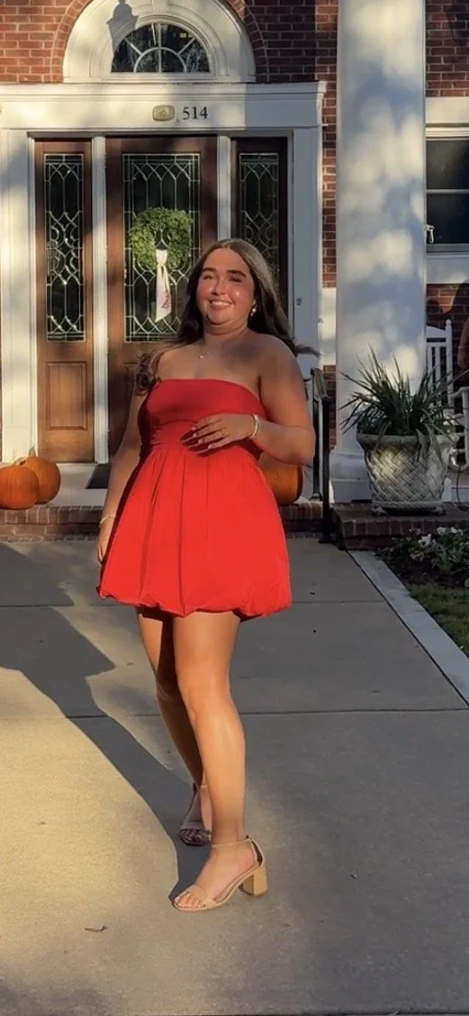 A smiling woman in a red strapless dress and beige high heels standing outside in front of a brick house with a wooden door, pumpkins, and potted plants.