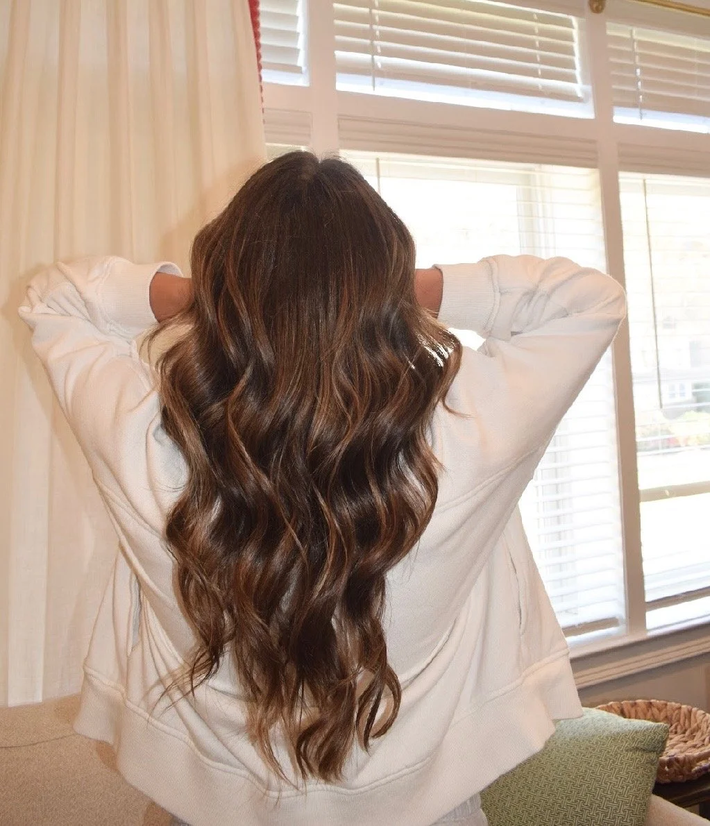 A woman with long, wavy brown hair standing in a room, facing away from the camera, with her arms raised behind her head, in front of a window with blinds.