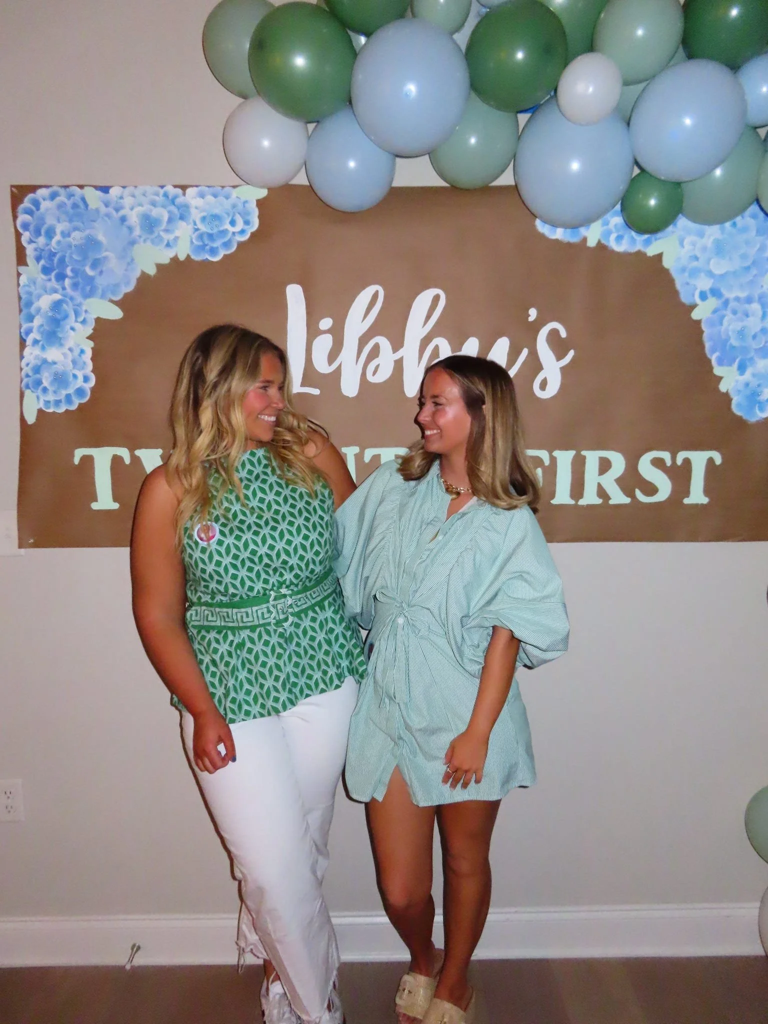 Two women smiling and standing close to each other in front of a brown backdrop with balloons and the words 'Libby's first time' printed on it. One woman is wearing a green geometric print top with white pants, and the other is wearing a light blue and white striped shirt dress.