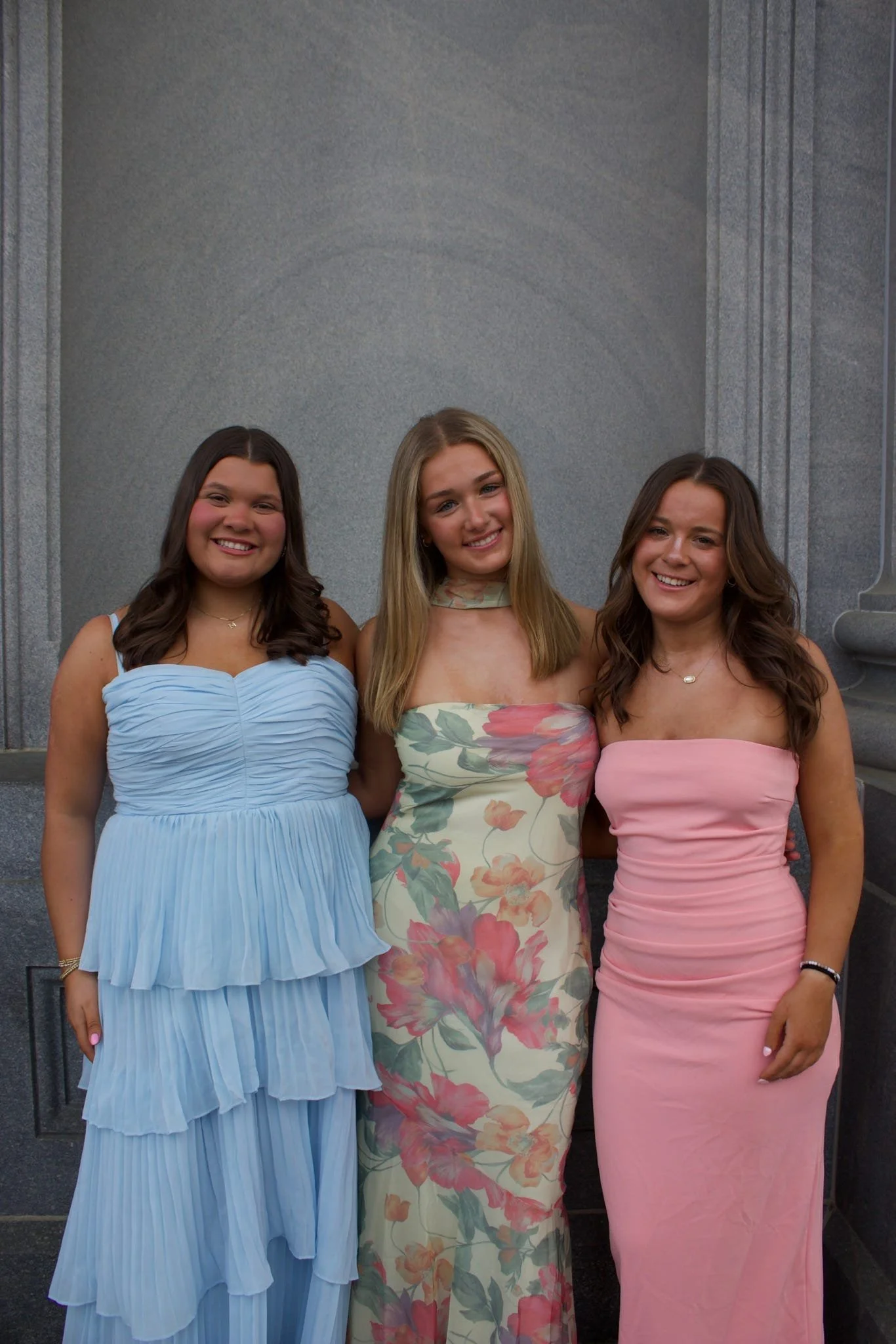 Three young women in formal dresses standing in front of a gray wall, smiling at the camera.