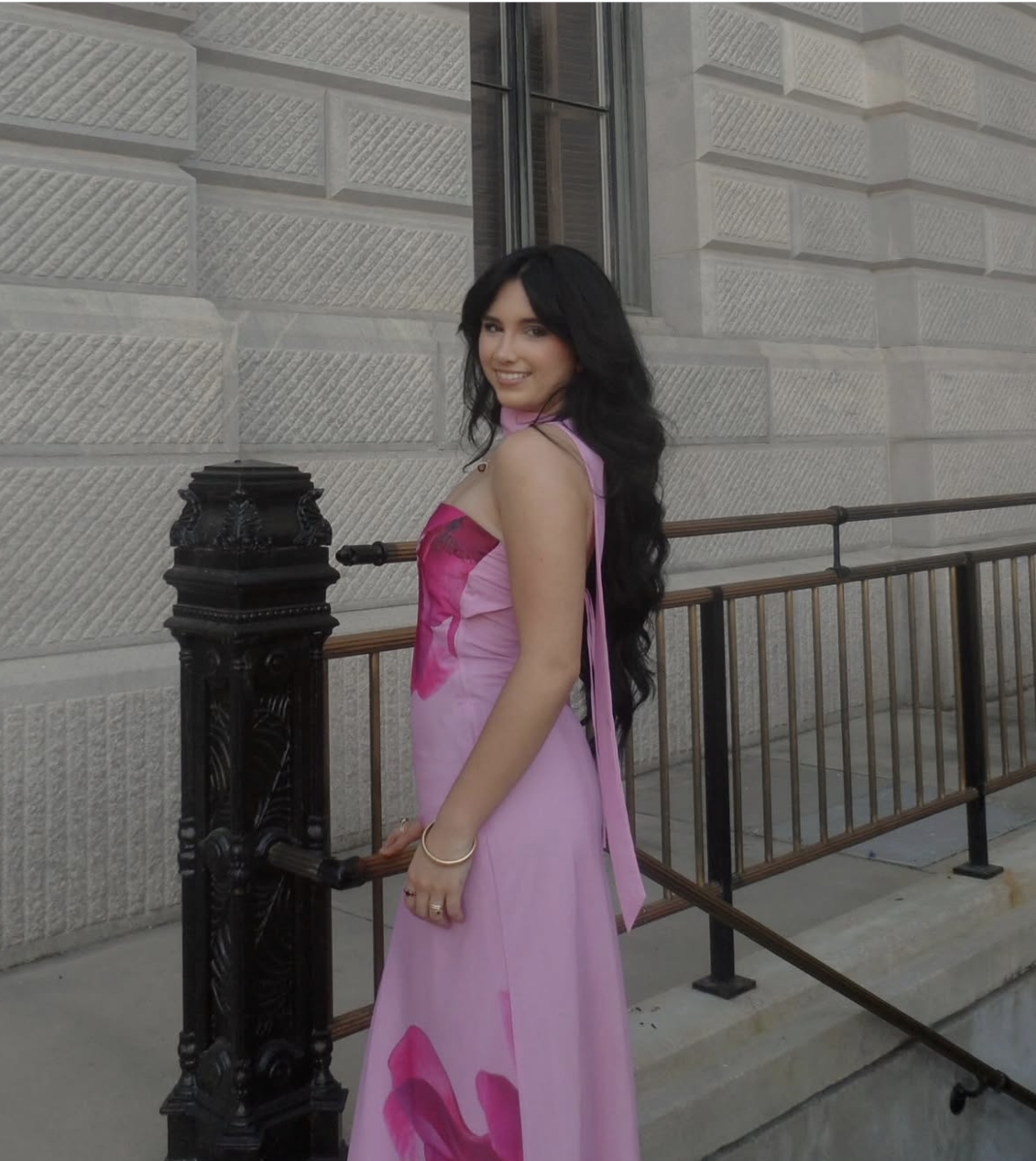 A woman with long black hair and a pink dress standing outside a stone building with black railing and a window.