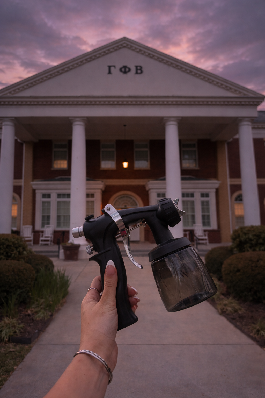 A person holding a spray bottle in front of a large, brick college or university building with Greek letters and columns, during sunset or sunrise.