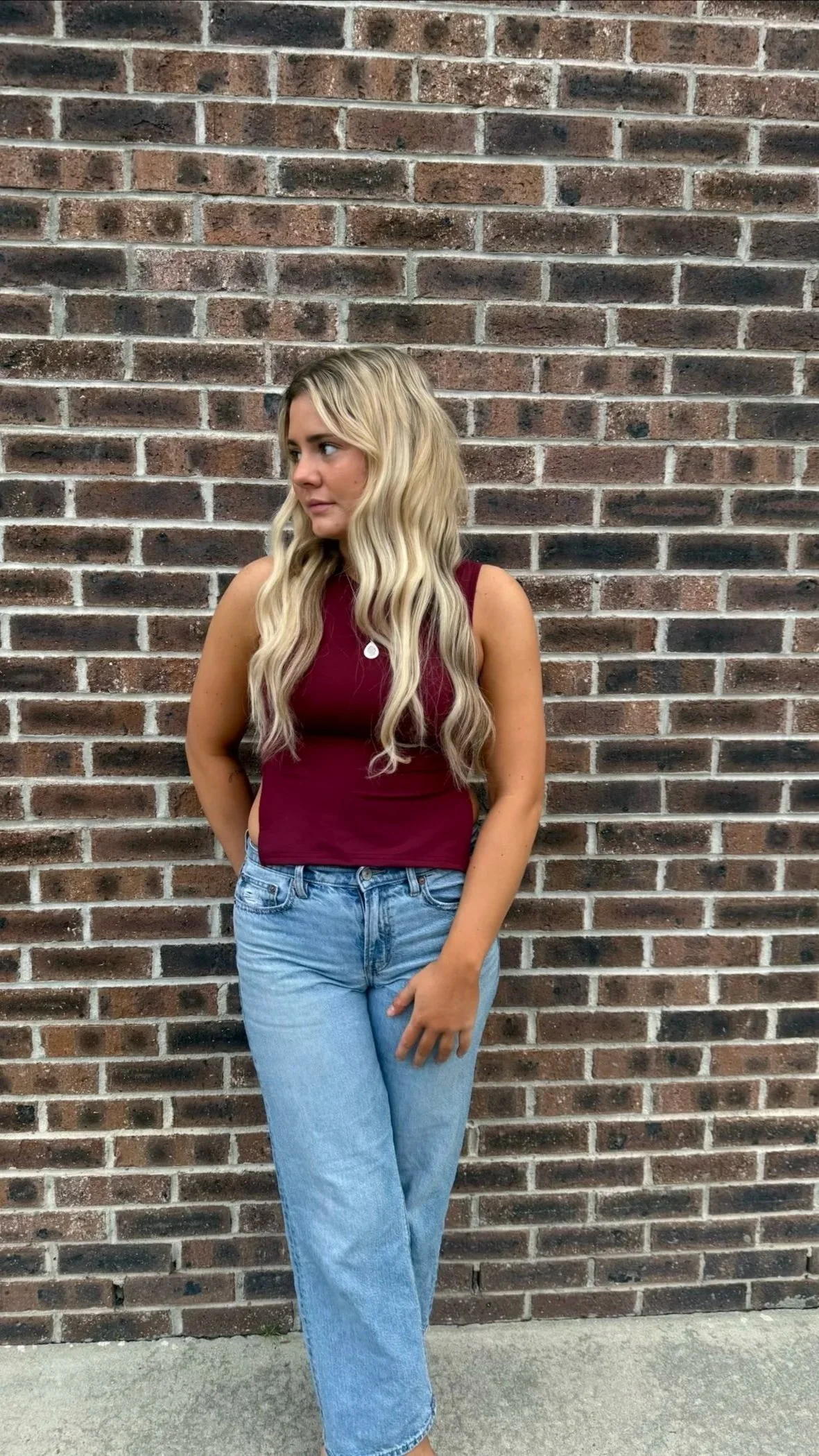 Young woman with long wavy blonde hair wearing a sleeveless maroon top and light blue jeans, standing against a brick wall.