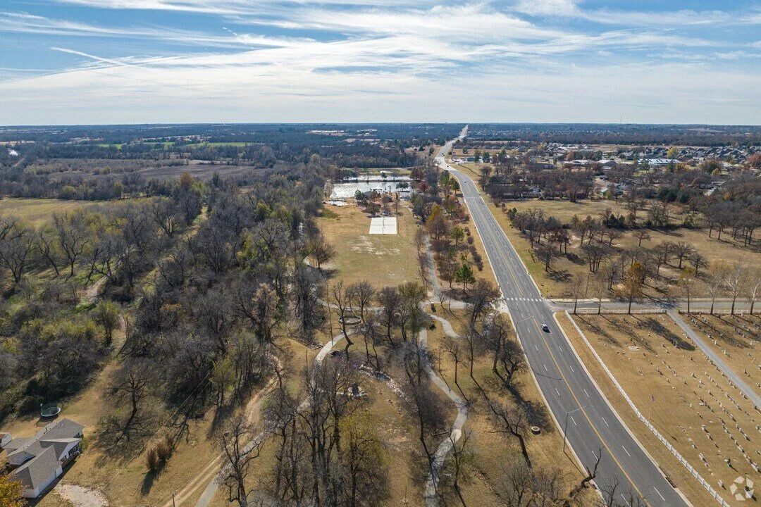 Aerial view of a rural landscape with a highway running through it, surrounded by trees, open fields, and bodies of water in the distance under a partly cloudy sky.