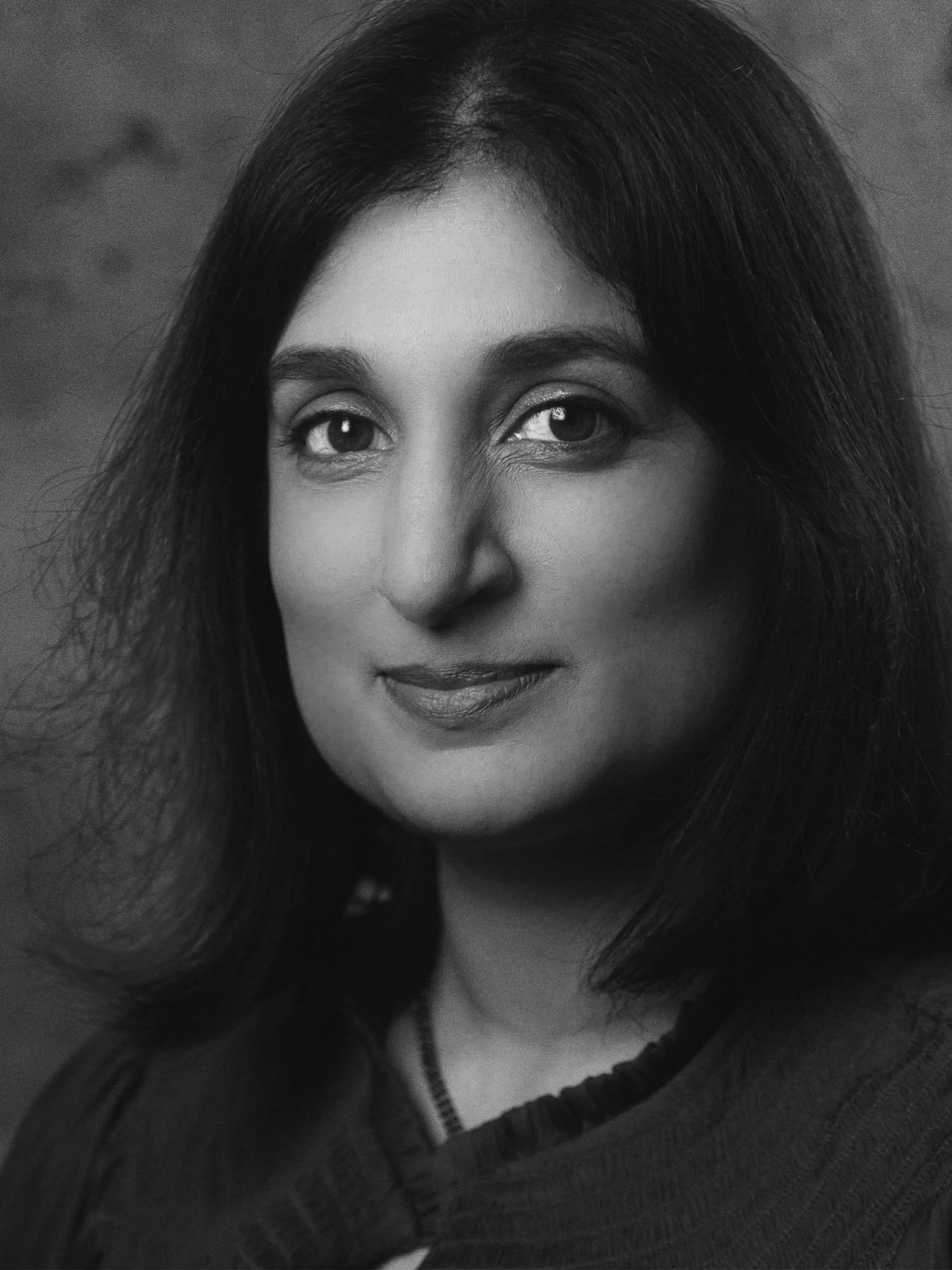 Black and white close-up portrait of Kavita Tomlinson with a subtle smile, wearing a black shirt.