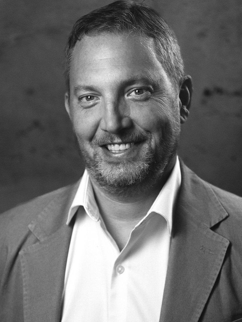 Black and white portrait of a smiling Andrew DeSio, wearing a suit jacket and collared shirt.