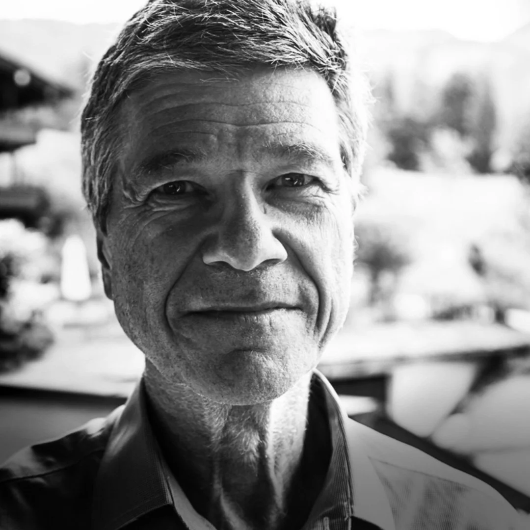 Close-up black and white photo of a smiling Jeffrey Sachs outdoors with blurred background, wearing a collared shirt.