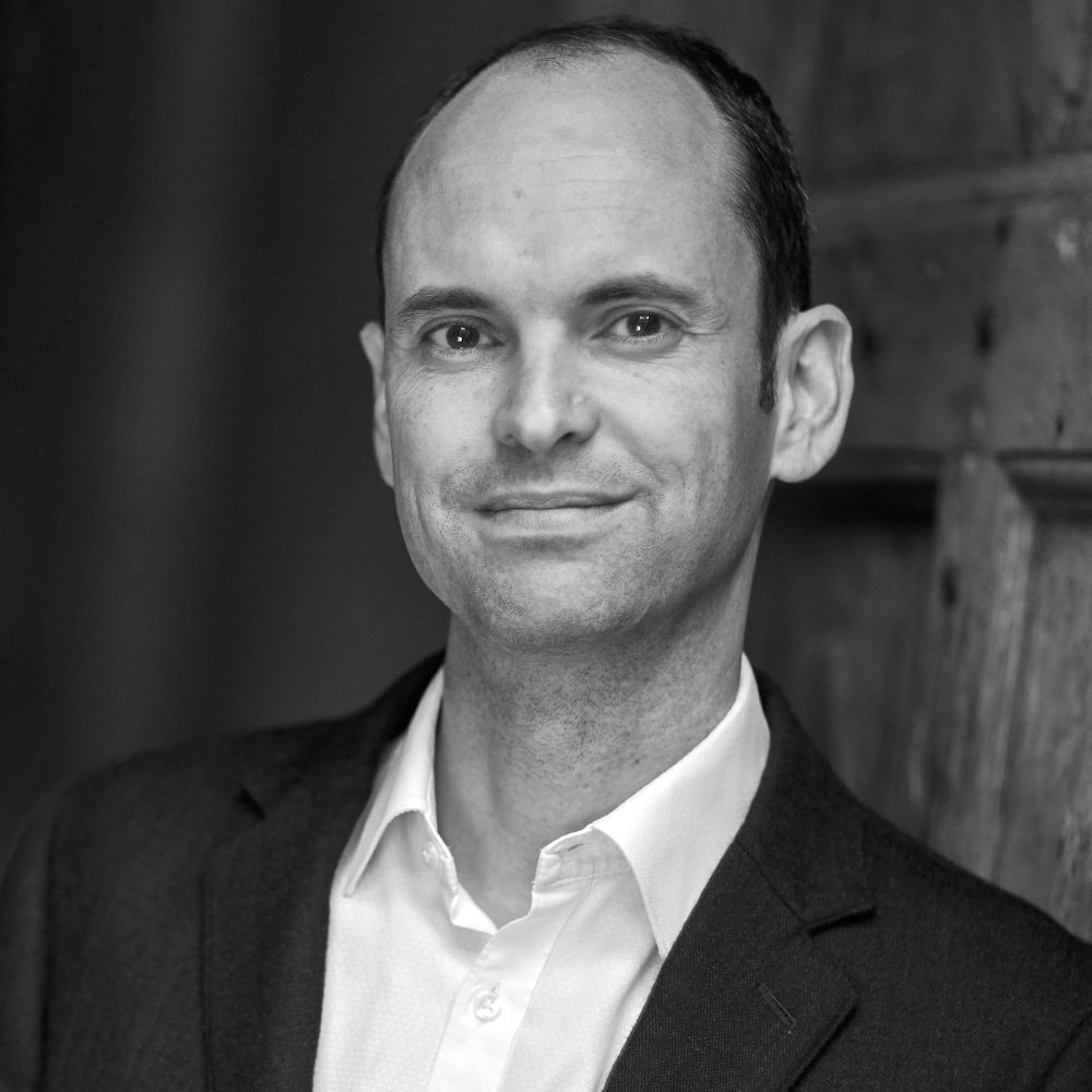 Black and white professional headshot of Toby Ord in a suit with a slight smile, standing in front of a wooden background.