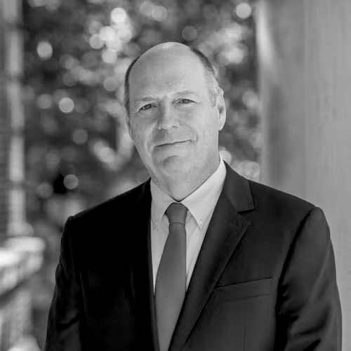 Black and white photo of Martin Schmidt in a suit and tie, standing outdoors with blurred trees in the background.