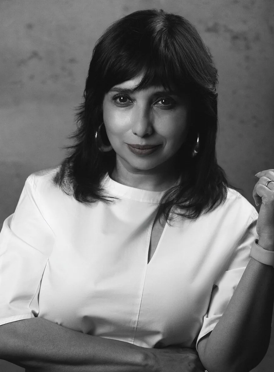 Black and white photo of Rimjhim Dey, wearing hoop earrings and a white top, sitting with her arm folded and looking into the camera.