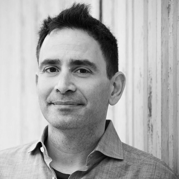 Black-and-white portrait of Elad Gil wearing a collared shirt, standing in front of a wooden wall.