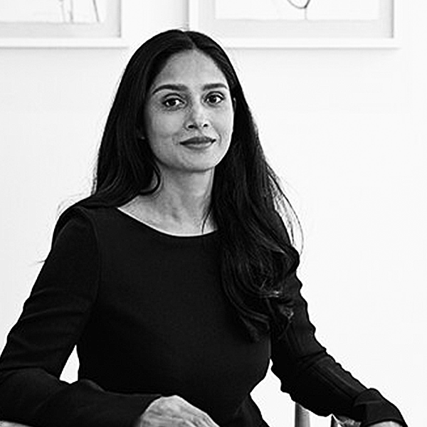 Saadia Madsbjerg, wearing a black top, sitting and looking at the camera against a background with framed pictures.