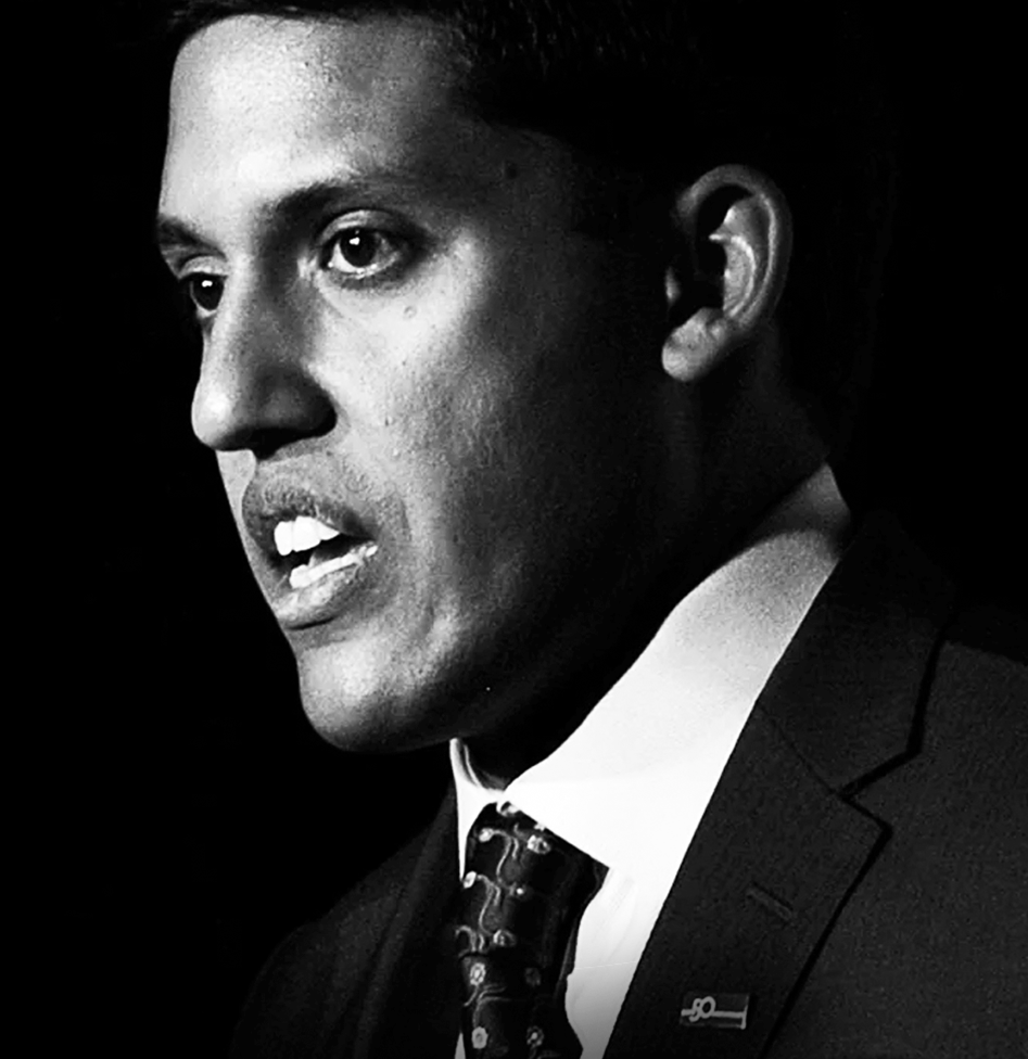 A black-and-white photograph of Dr. Rajiv Shah in formal attire, with a serious expression, short hair, and an earpiece, giving a speech.