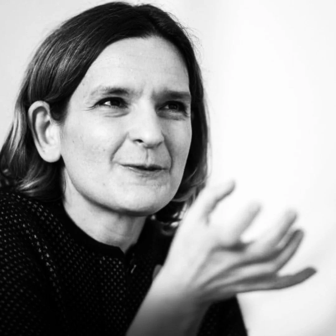 Black and white photo of Esther Duflo, smiling slightly, with her hand raised.