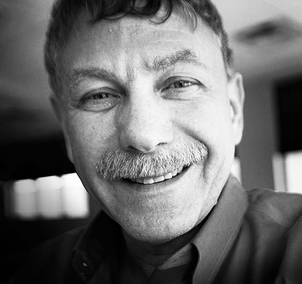 Close-up black and white photo of a smiling Eric Lander with a mustache, wearing a collared shirt.